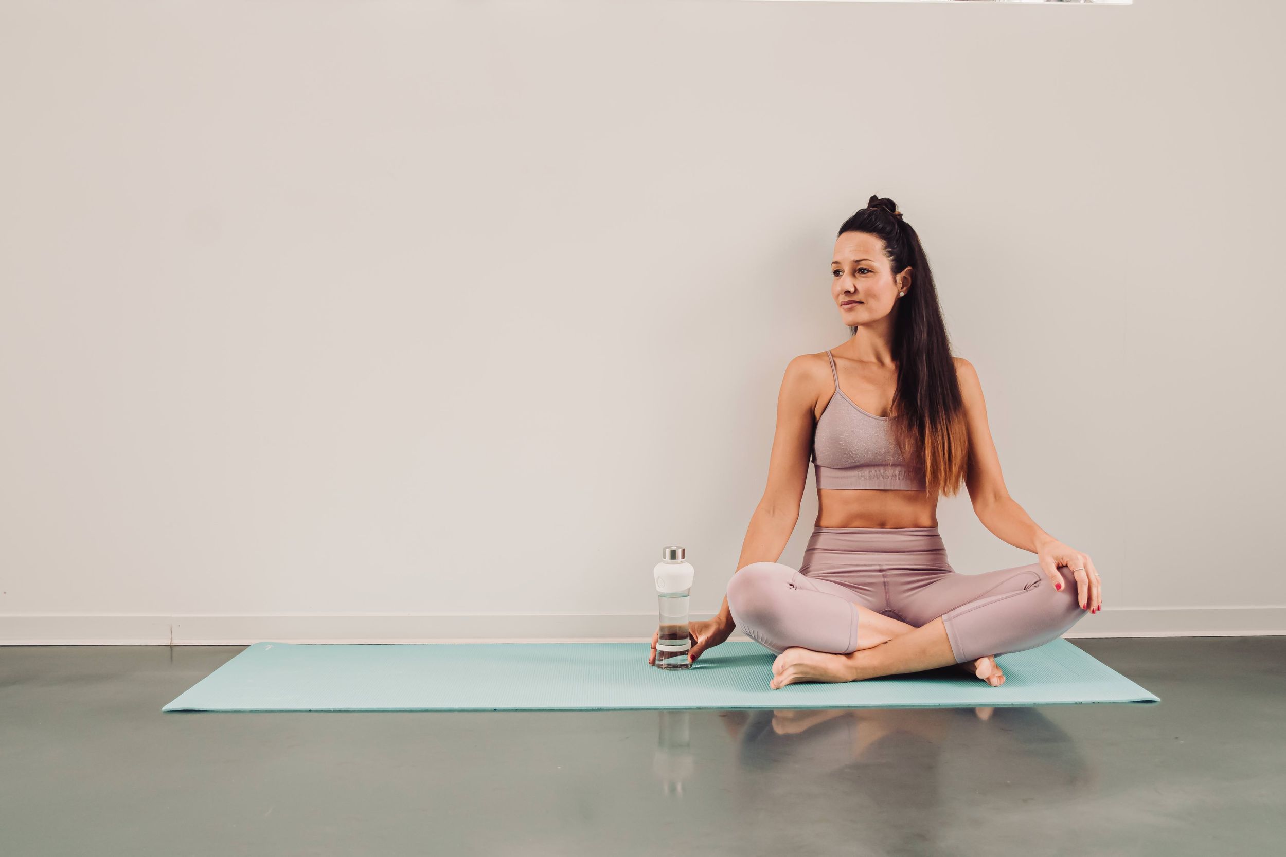 Person in workout attire sits on a yoga mat while holding a smartphone during an indoor exercise session.