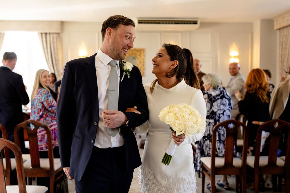Wedding guests gather in a reception hall with wooden chairs and white walls.