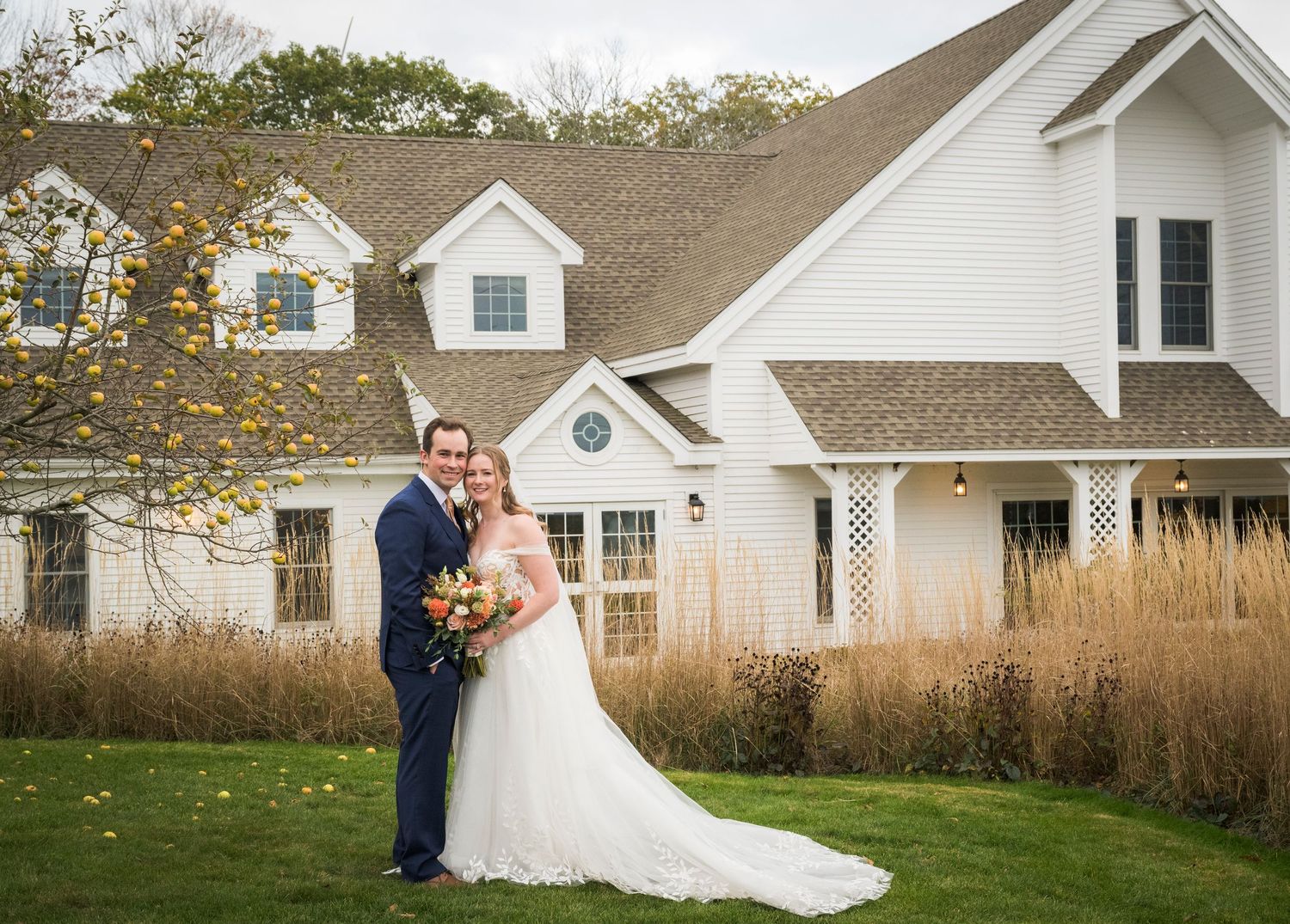 A wedding couple poses in front of a white farmhouse with tall grass and autumn trees in the background.