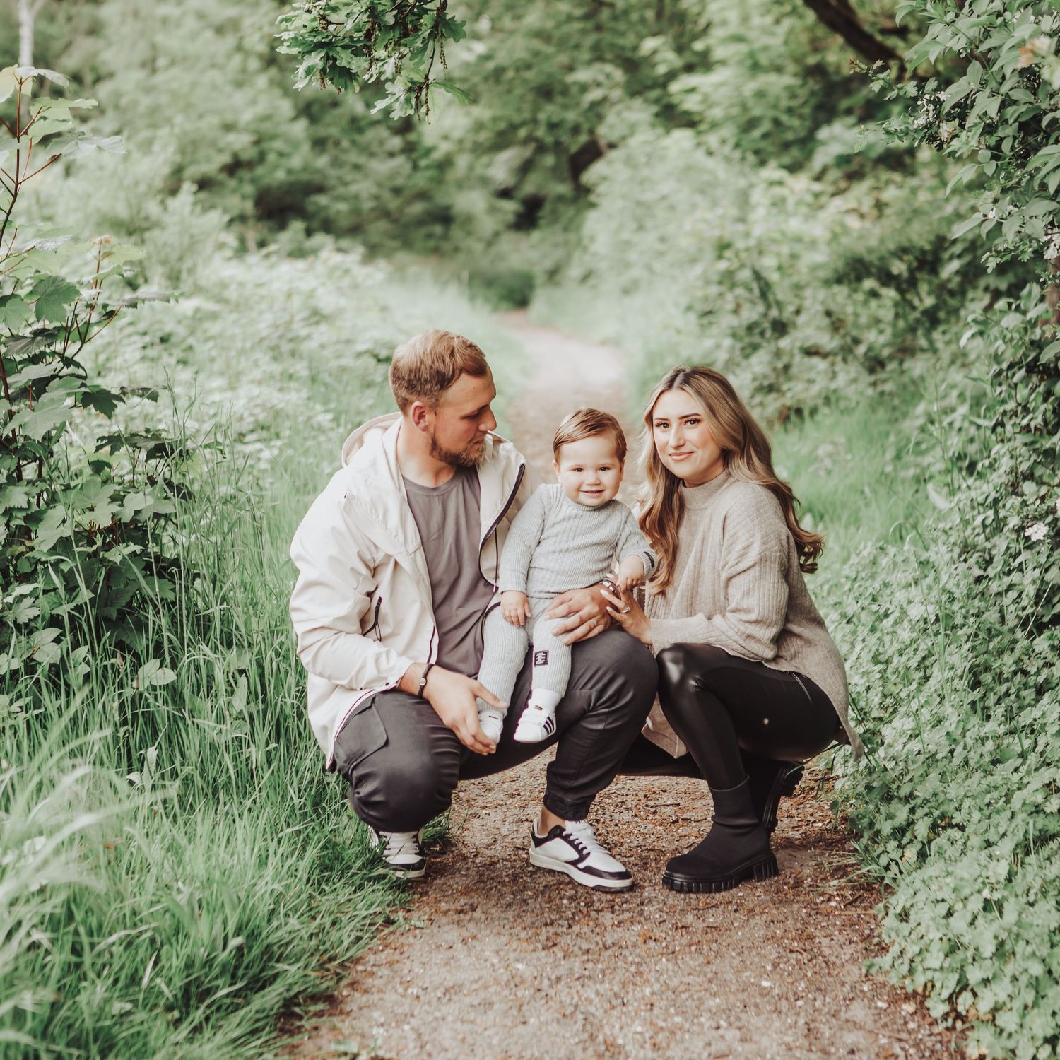 Family of 3 on a woodland path, crouching down holding their little boy - smiling to the camera.