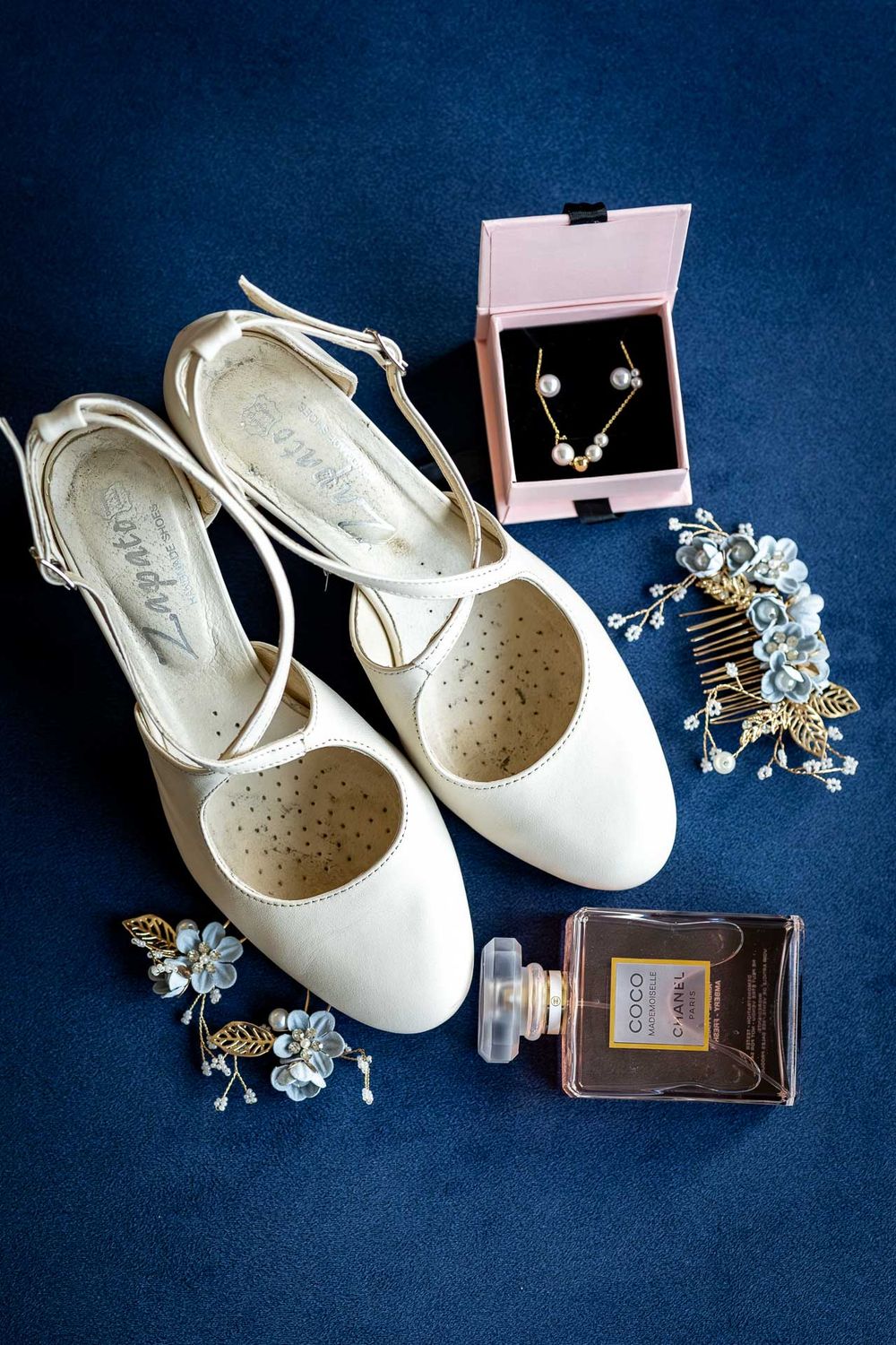 White bridal flats with jewelry and accessories arranged on navy blue background.