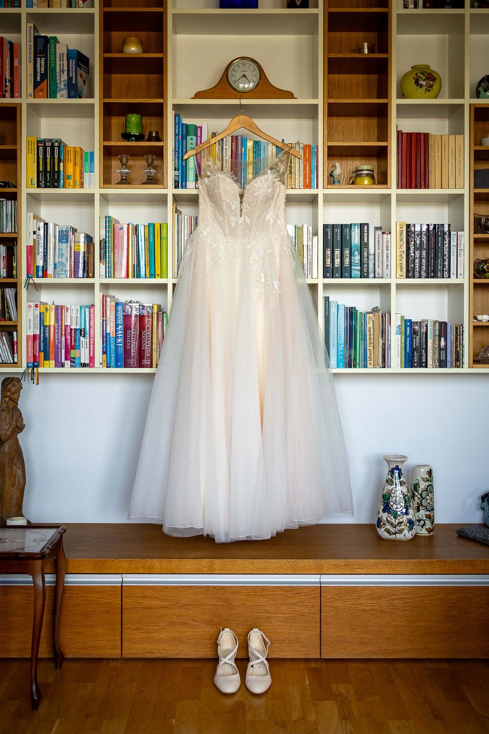 White wedding dress displayed against built-in bookshelves with shoes beneath.