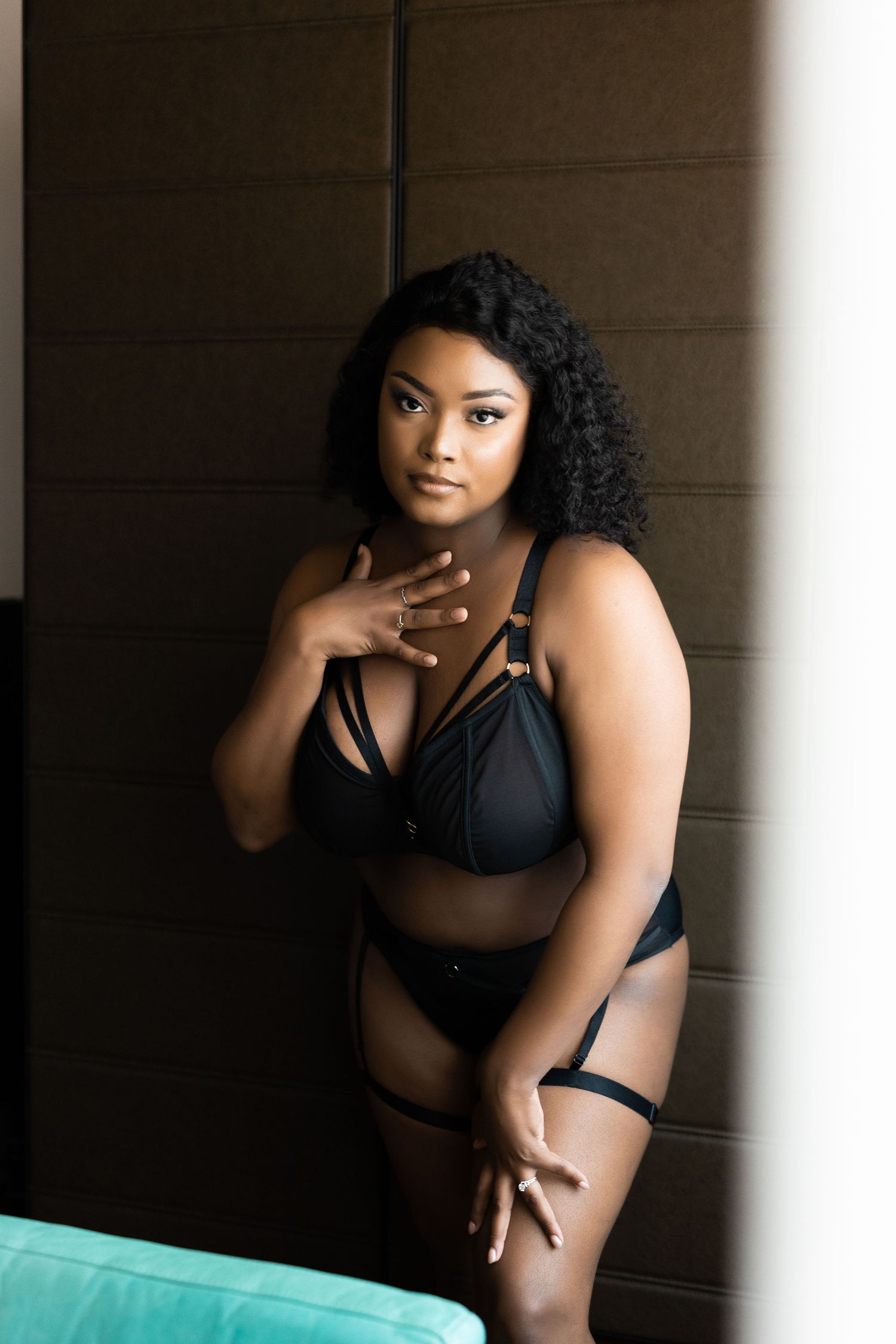 Curvy model poses confidently in black lingerie against brown wall.