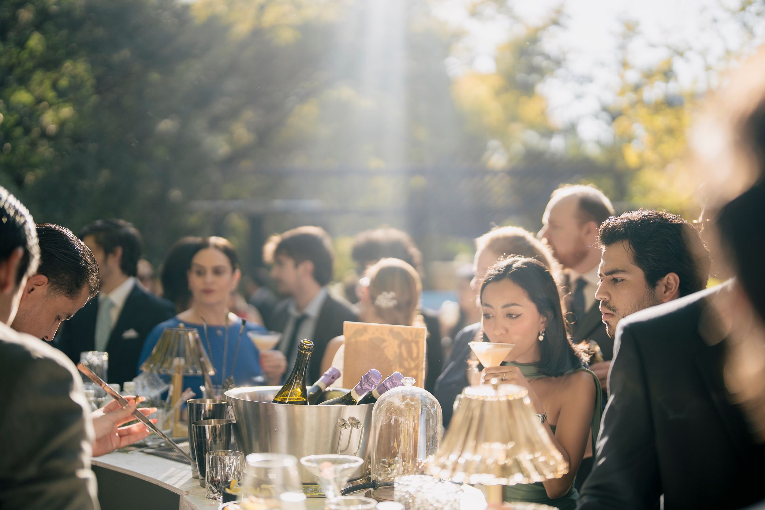 Wedding guests enjoy an outdoor reception dinner at long tables under golden sunlight in a garden setting.