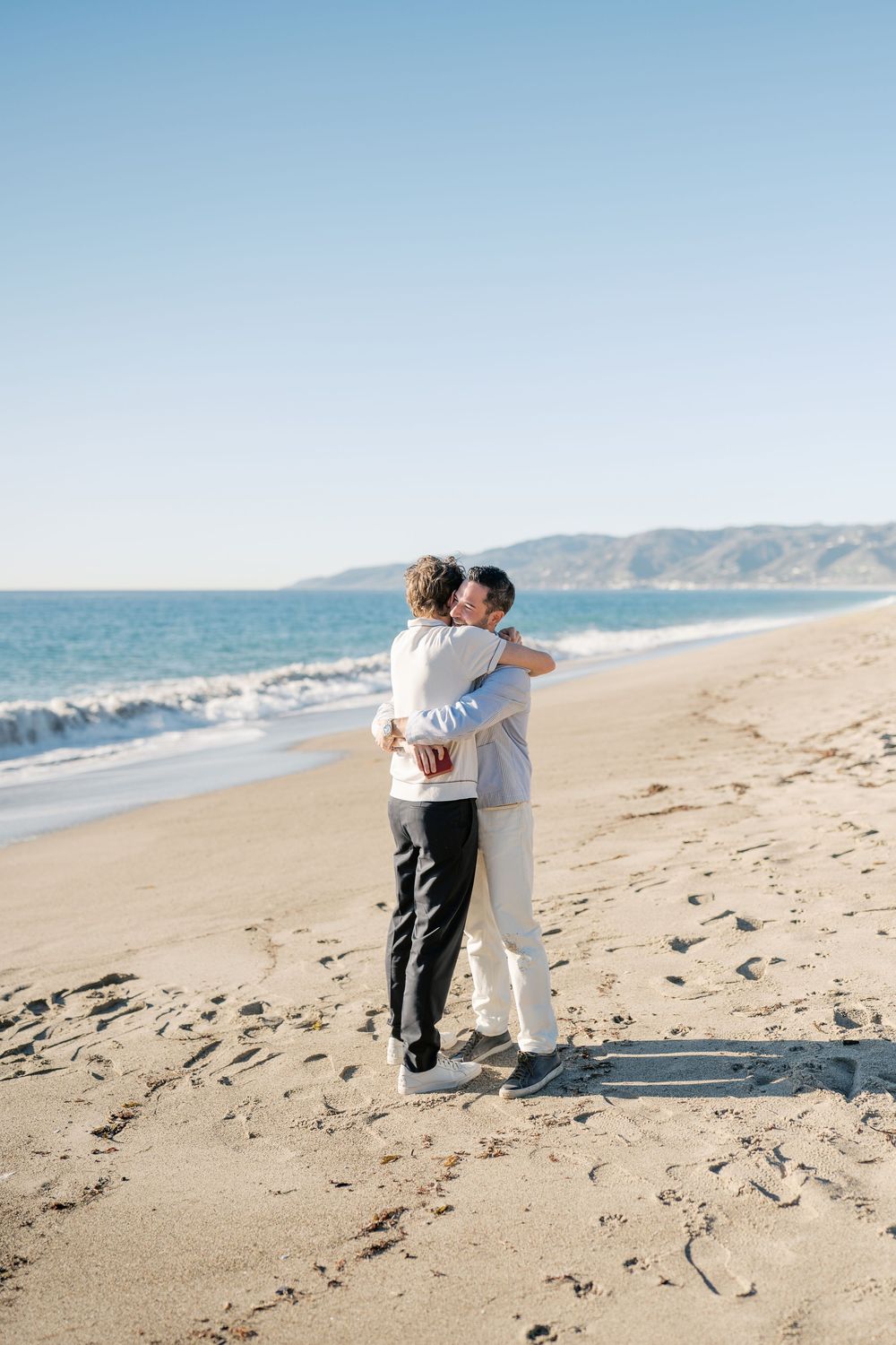 Two people share an intimate embrace while walking along a scenic beach with mountains in the background.