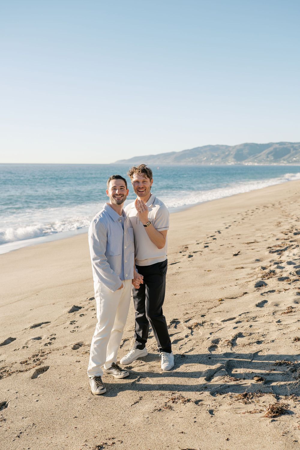 Two people in contrasting outfits walk together along a sandy beach with mountains in background.