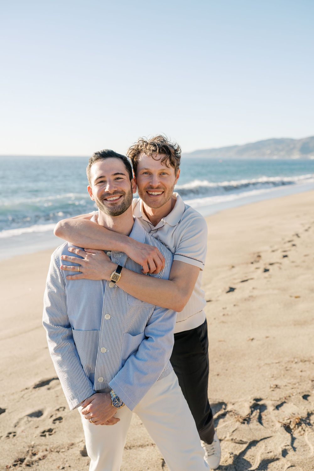 Two people share a joyful embrace on a sunny beach with ocean waves and mountains in the distance.