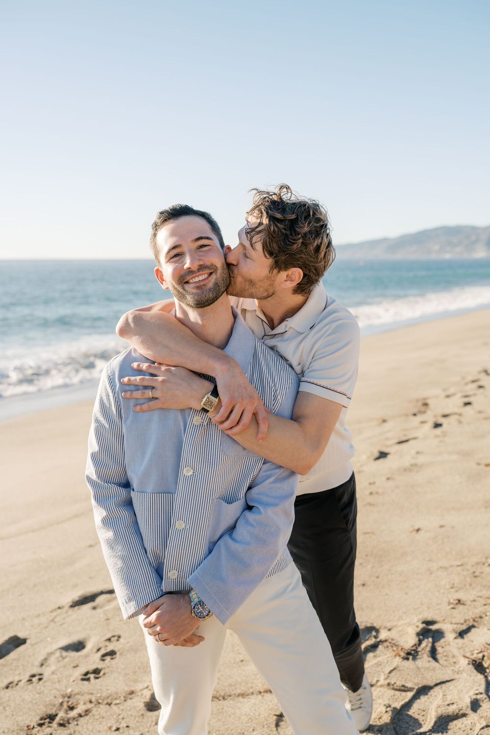 A couple shares playful moments on a scenic beach with ocean waves in the background.