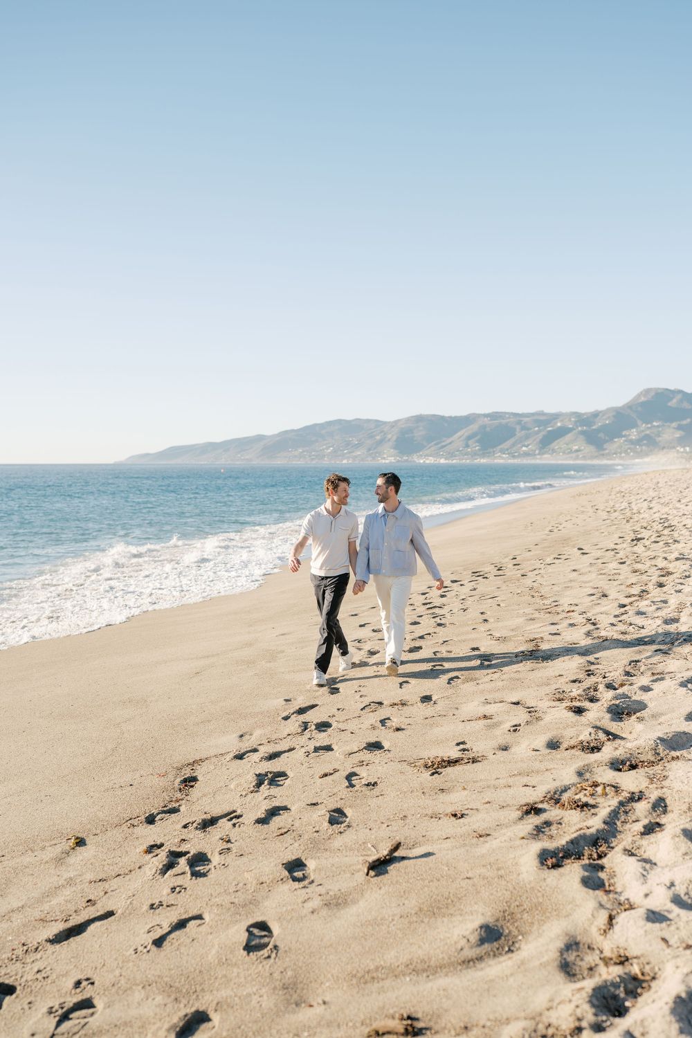 Two people in white walking along a scenic beach with mountains in the background.