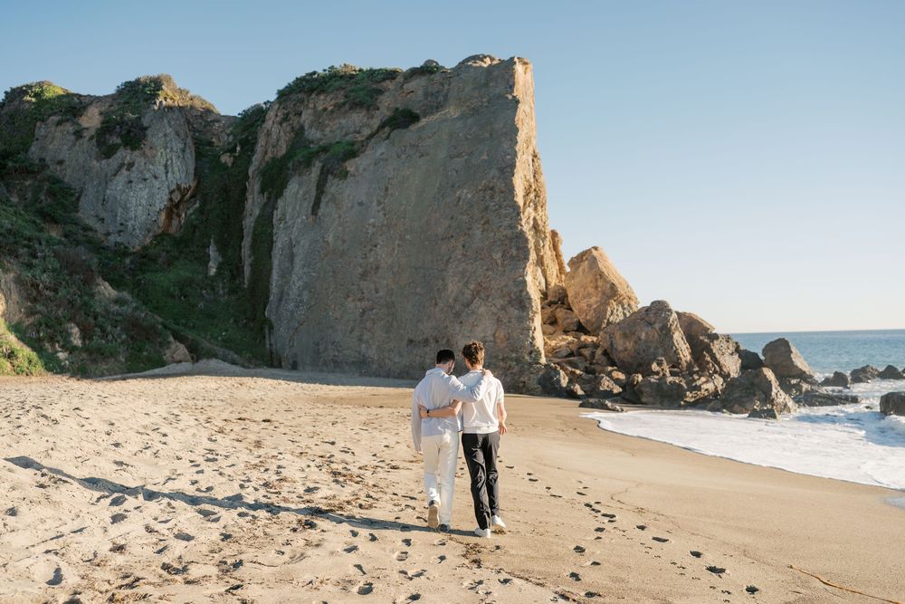 A couple walks hand in hand on a scenic beach with dramatic coastal cliffs in the background.