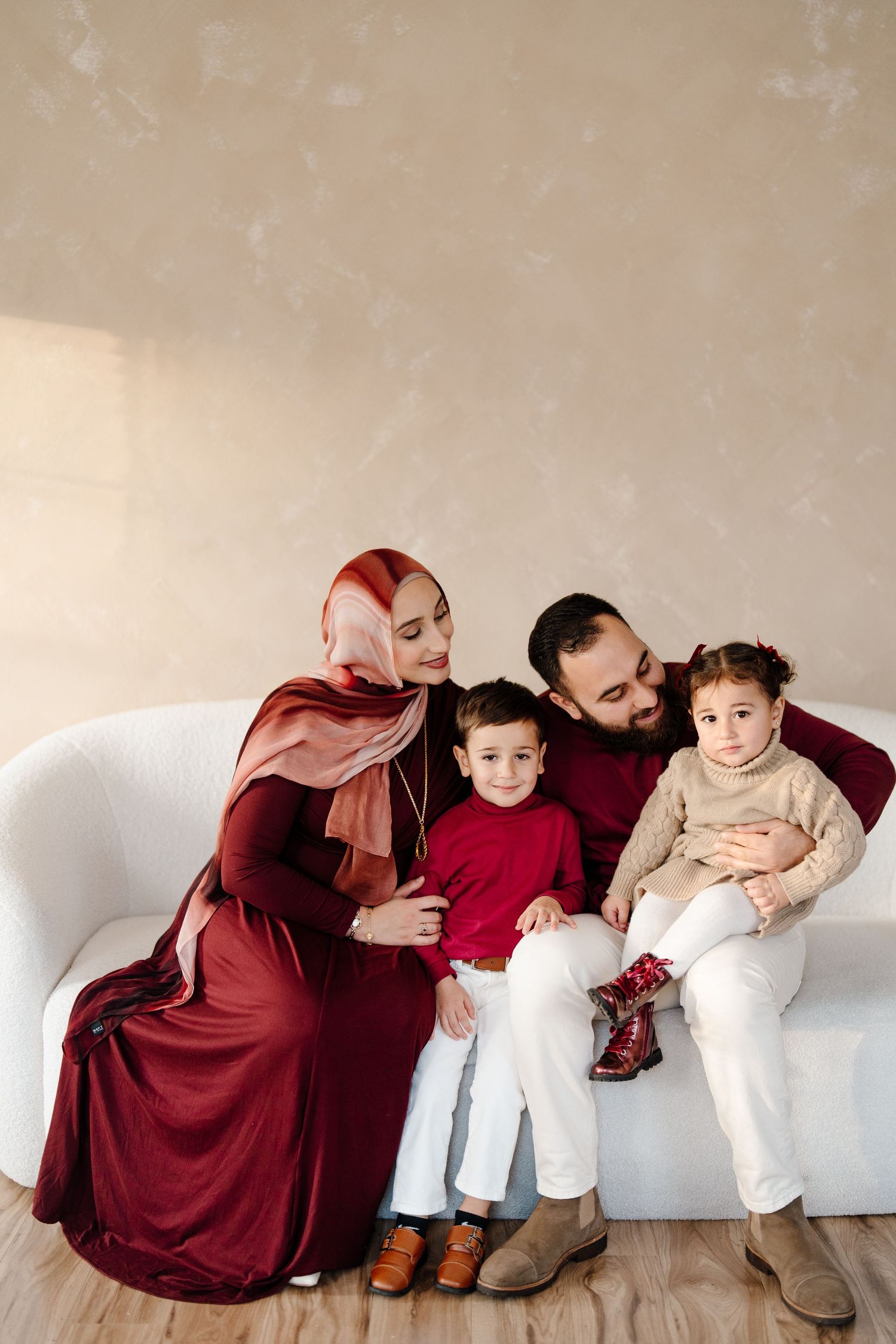 Muslim family of four sitting together on a white sofa, parents in burgundy outfits with mother wearing hijab.