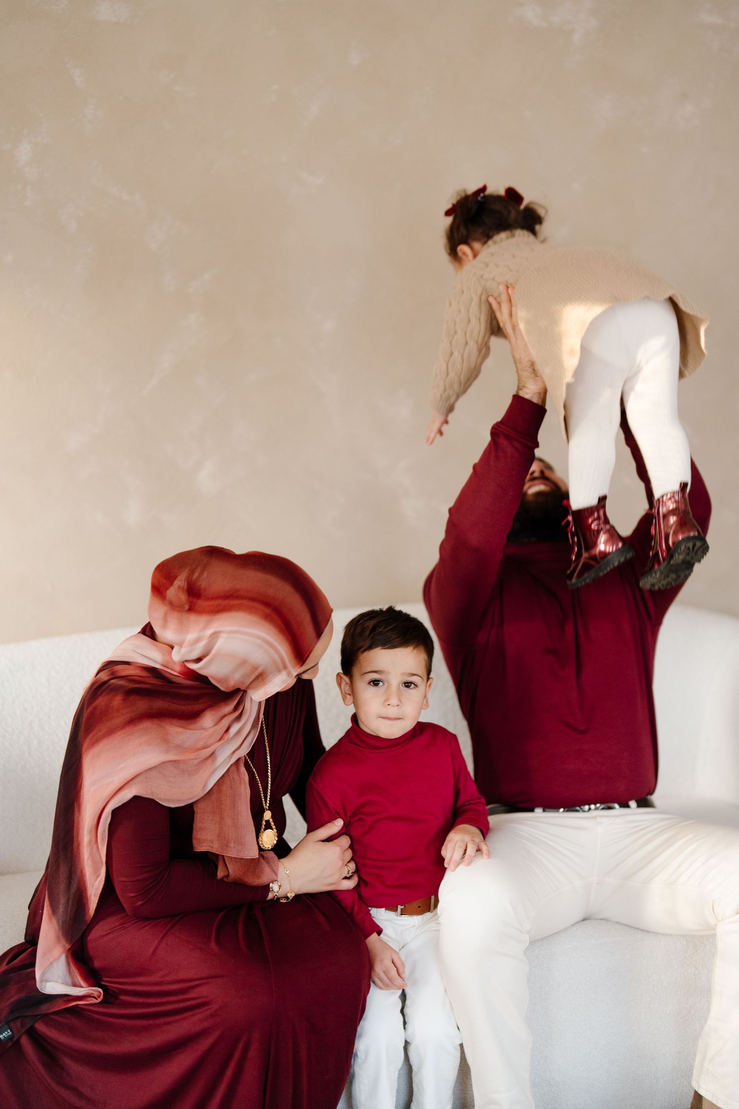 Muslim mother in burgundy hijab holds son while father lifts daughter playfully on a white sofa.