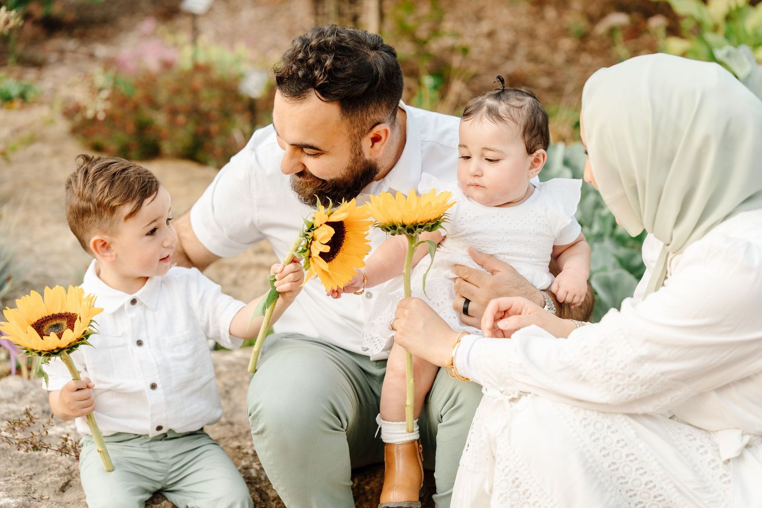 A Muslim family with two young children admires sunflowers together in a lush garden setting.
