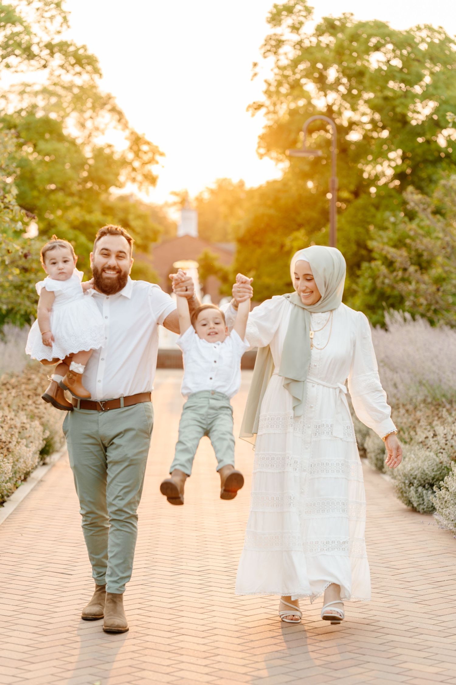 A joyful Muslim family swings their toddler between them while walking along a sunlit garden pathway.