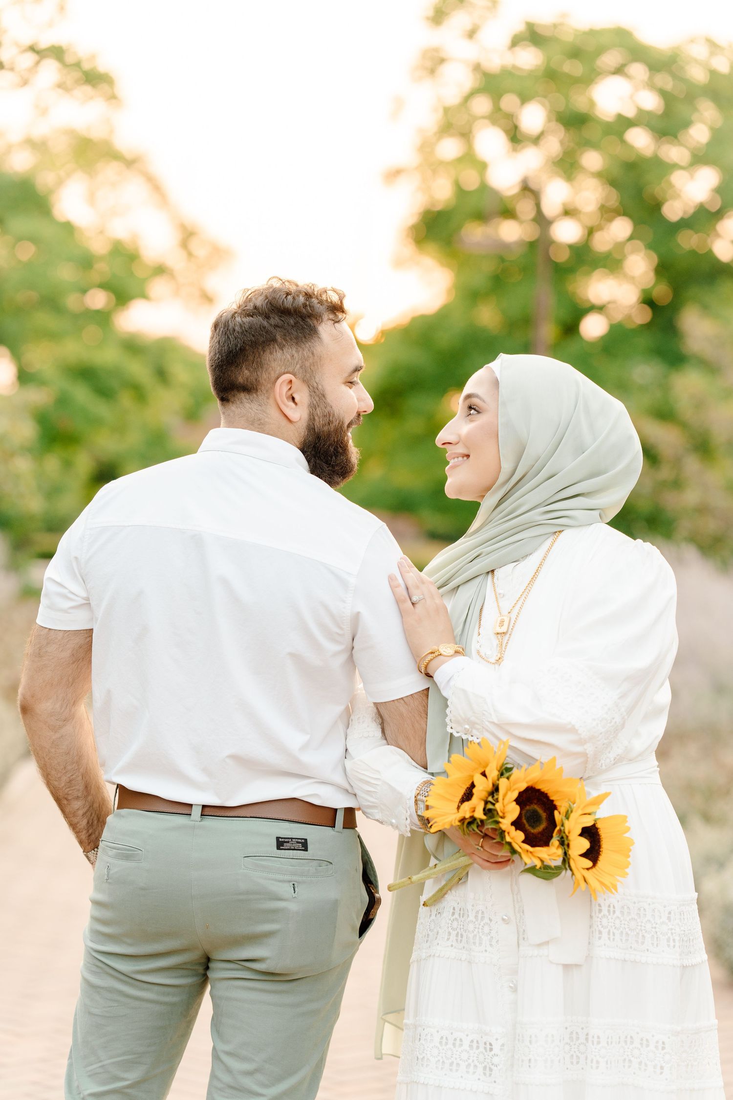 A Muslim couple in white and sage outfits shares a smile while holding sunflowers in a golden sunset garden.