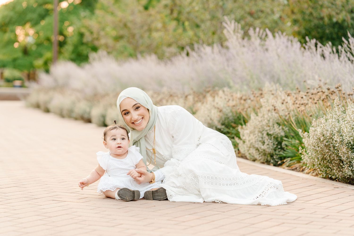 A Muslim mother in a white hijab and dress sits on a brick path smiling with her baby in a garden.