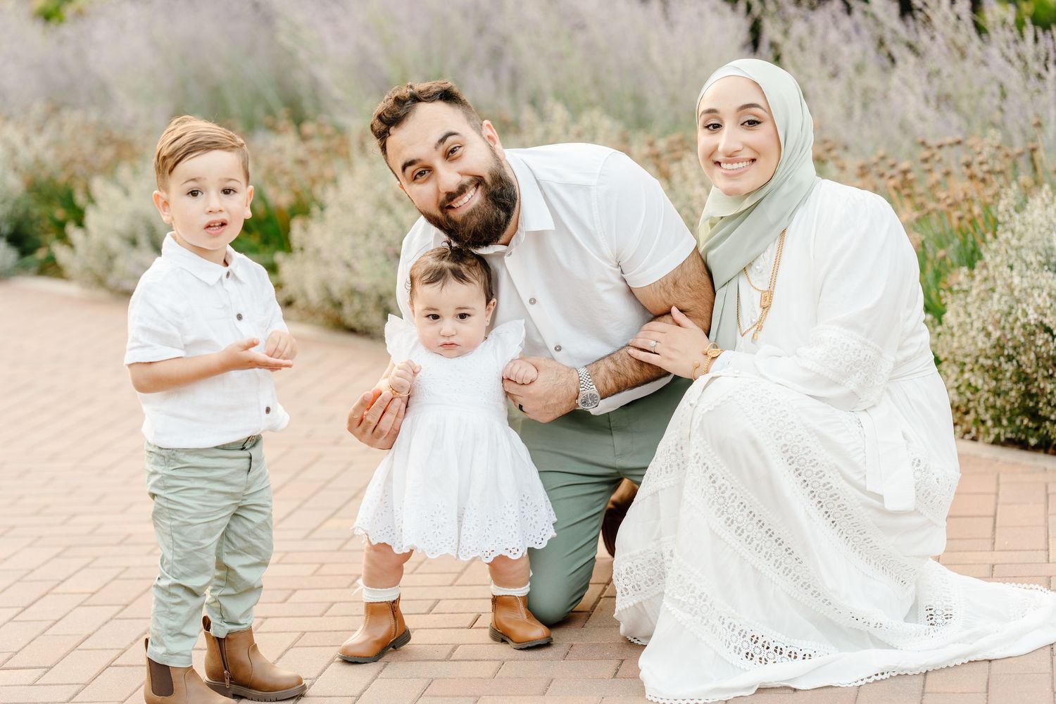 A smiling Muslim family of four poses together on a brick pathway surrounded by lavender in a garden.