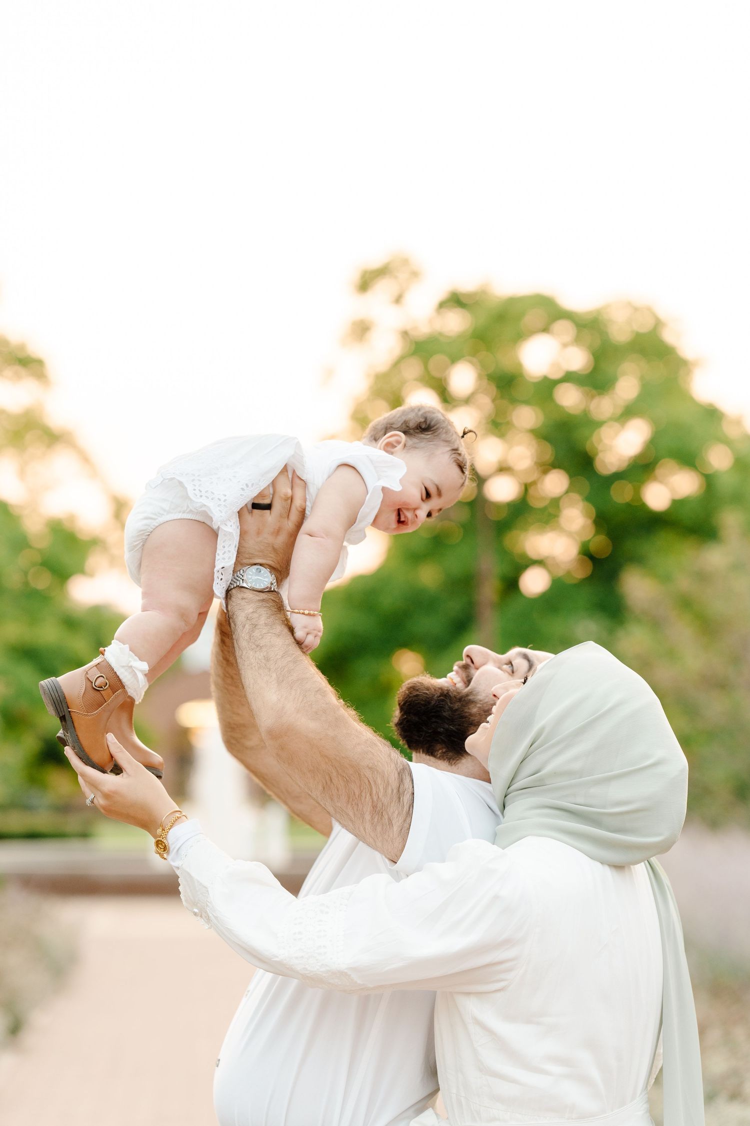 A bearded father in a white outfit joyfully lifts his laughing baby girl into the air outdoors.