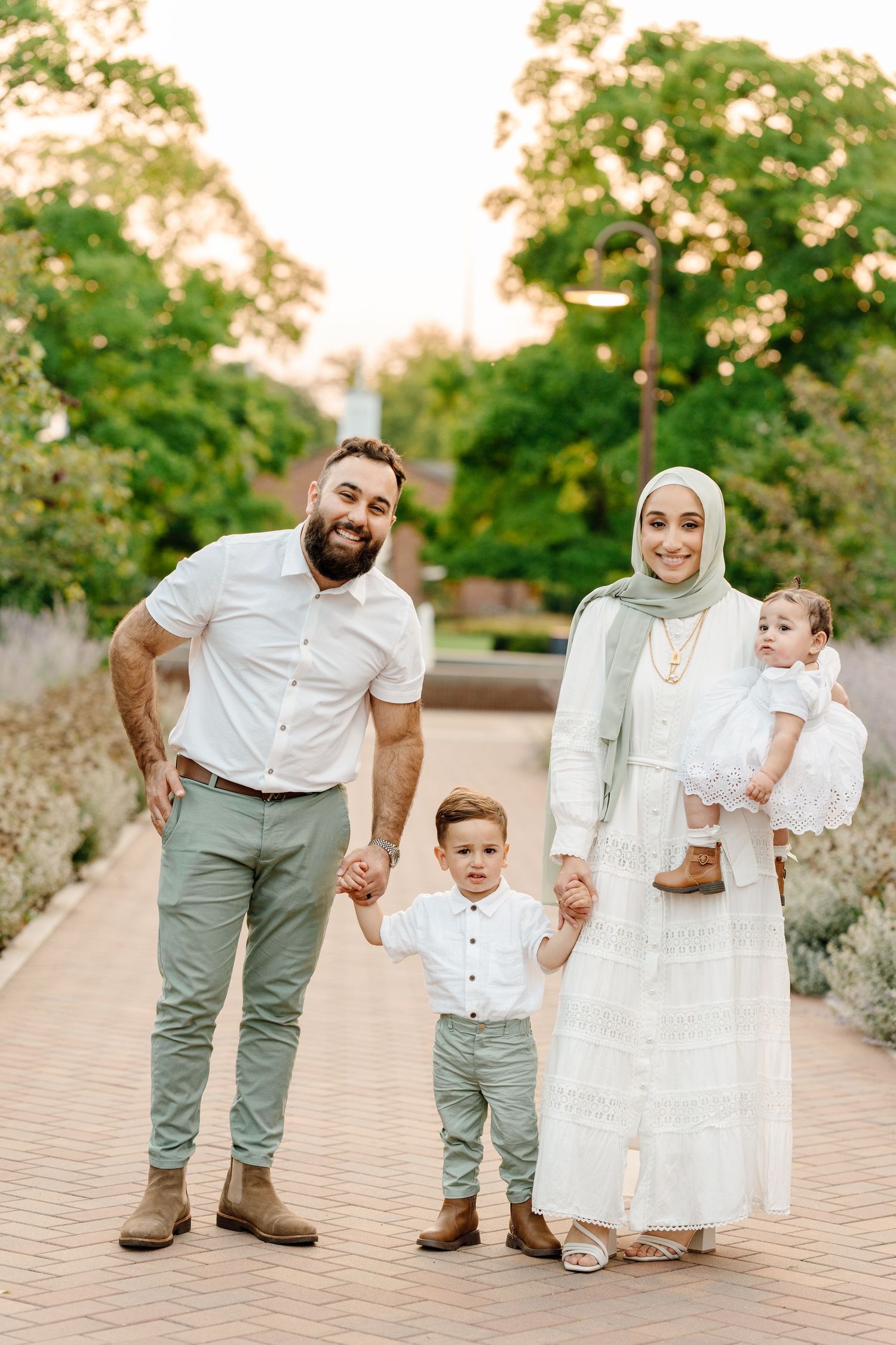 A Muslim family of four in coordinated white and sage outfits poses together on a sunny garden pathway.
