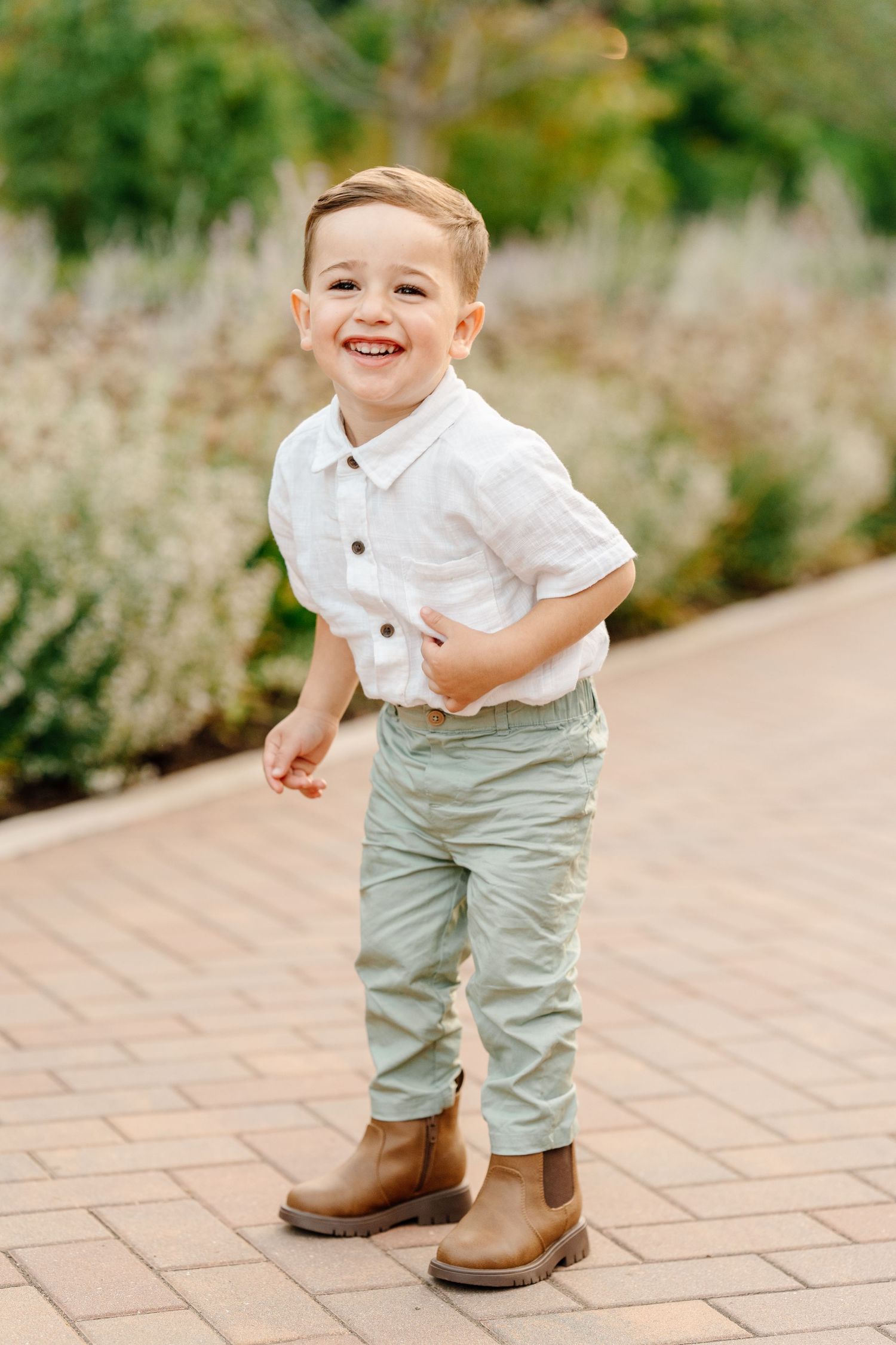 A cheerful young boy in a white shirt, sage pants, and brown boots smiles on a brick garden path.
