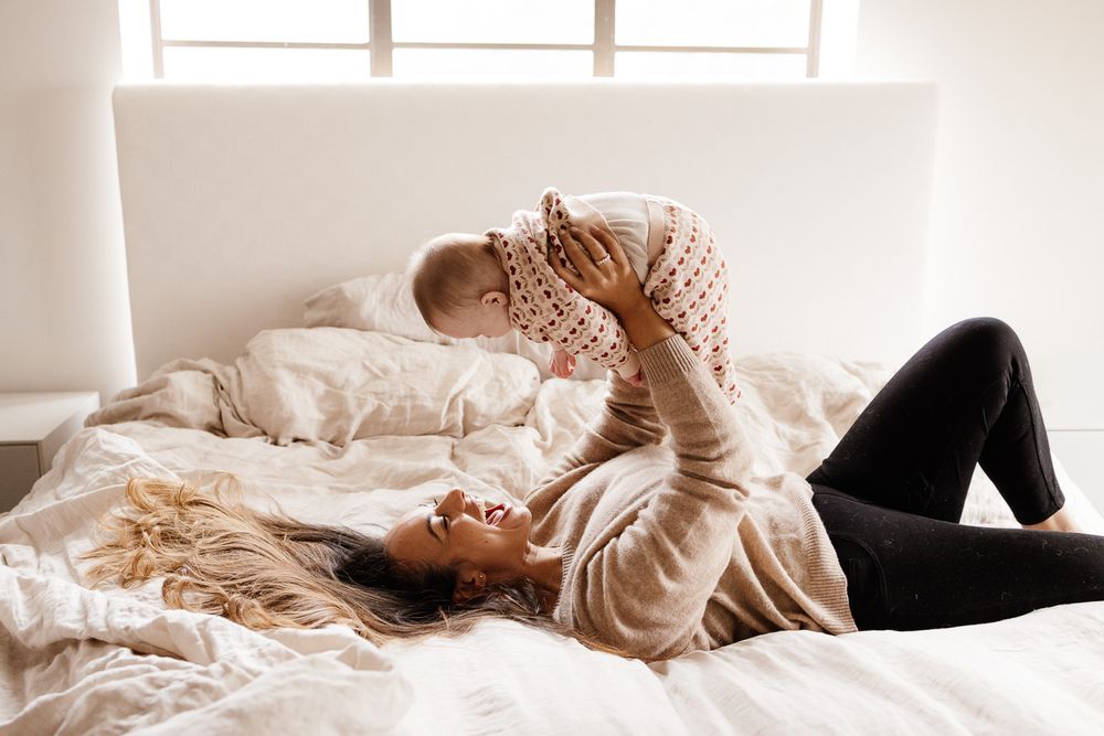 A mom holds her baby up in the air while laying on her bed at their in-home photo session in Los Gatos