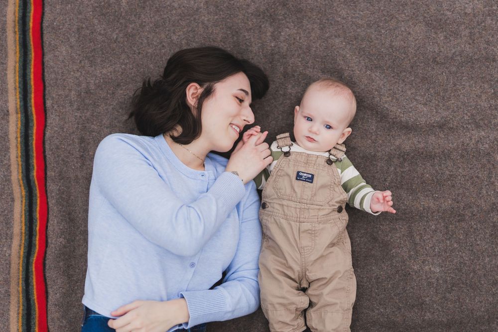 A parent and baby share tender moments while resting on a blanket during an outdoor photoshoot.