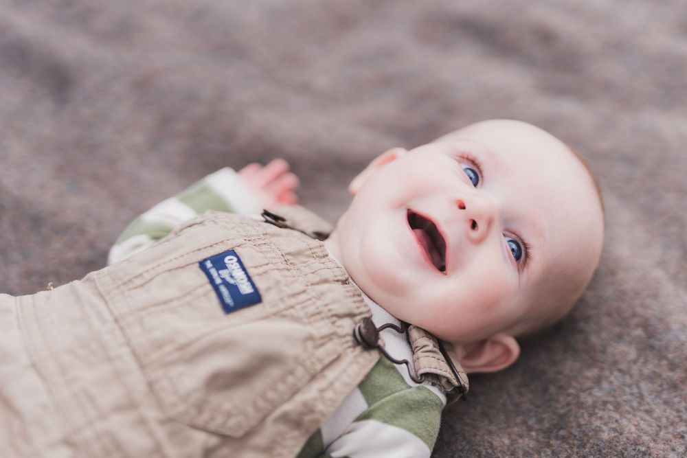 A baby lies on sandy ground while looking up at the sky with a curious expression.