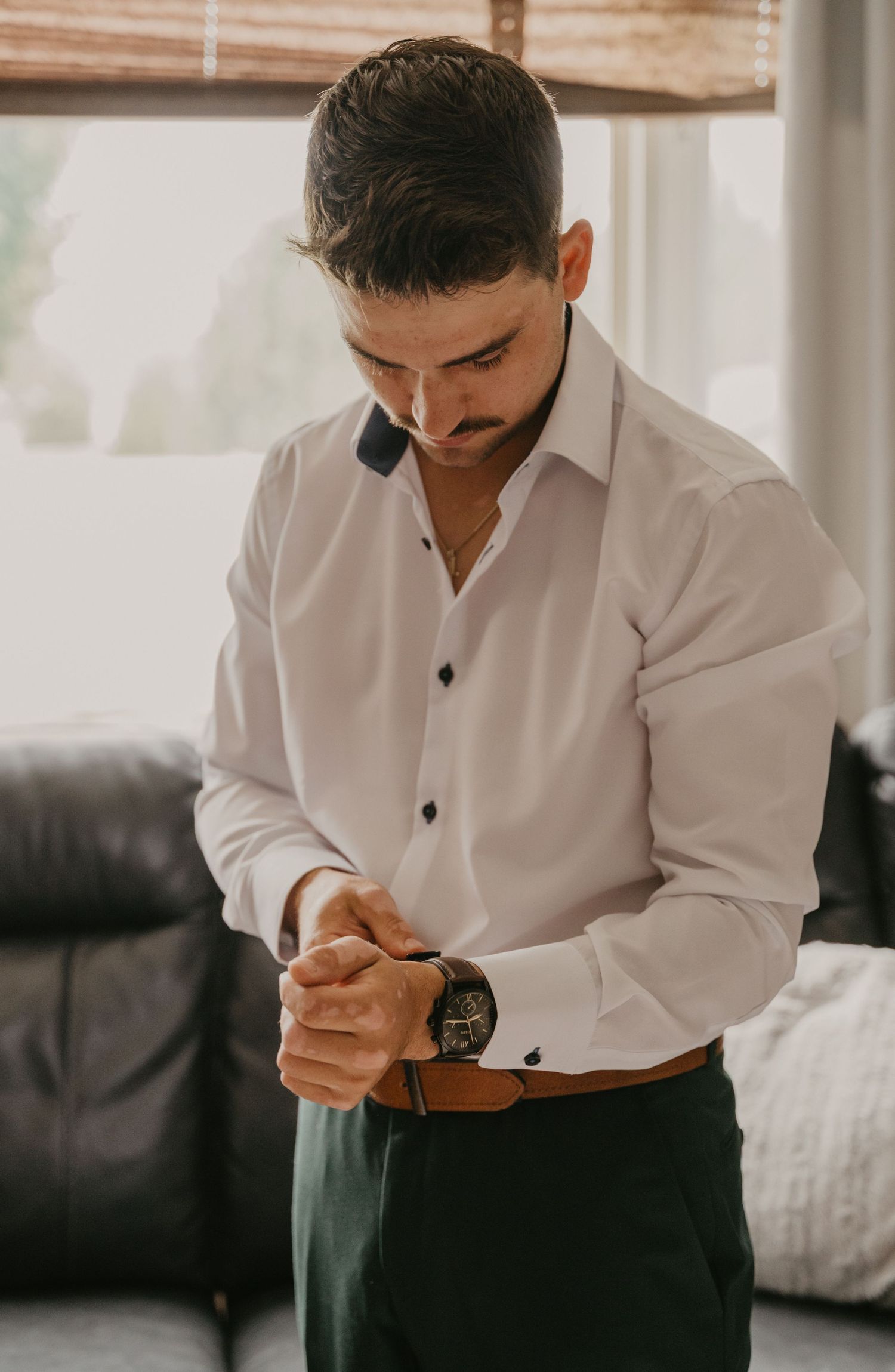 Groom in white dress shirt and black pants adjusting cufflinks while getting ready.