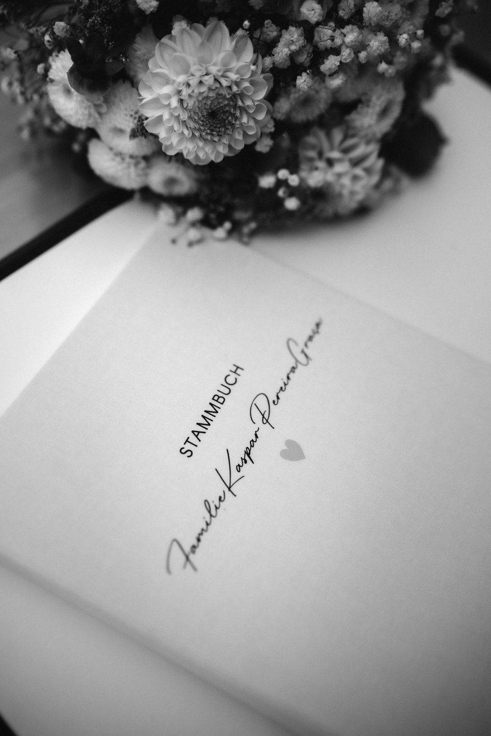 Black and white photo of a wedding guest book with handwritten signature and flowers in soft focus on top.