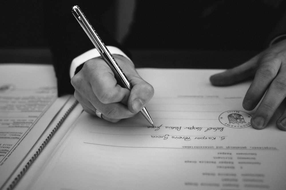 Black and white close-up of hands writing with a pen on paperwork or documents.