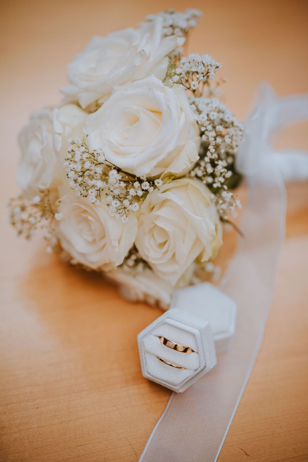 A white rose wedding bouquet with baby's breath lies beside a hexagonal ring box on a wooden surface.