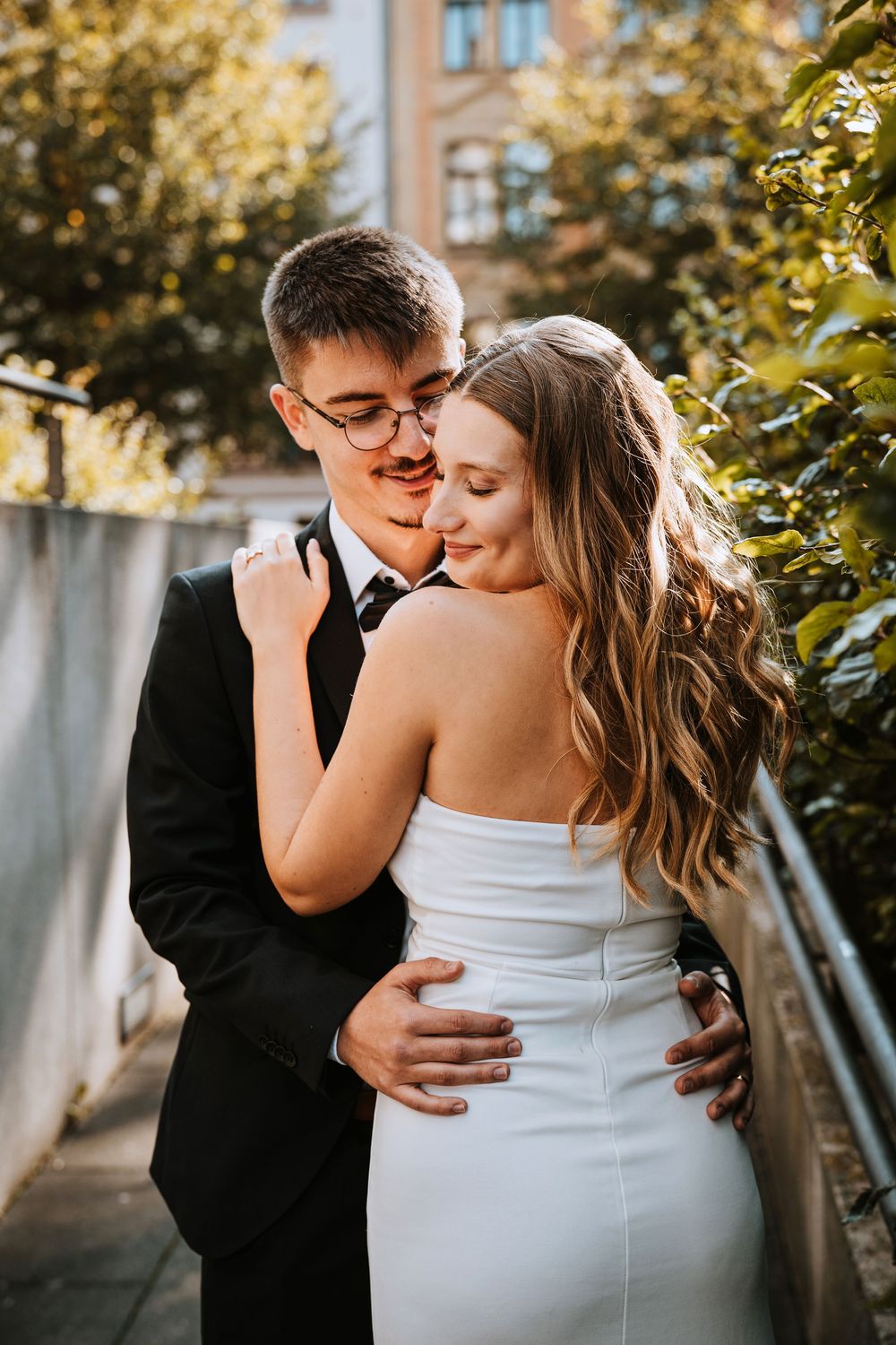A couple embraces intimately outdoors, with the bride in a white strapless gown and groom in classic black suit at sunset.