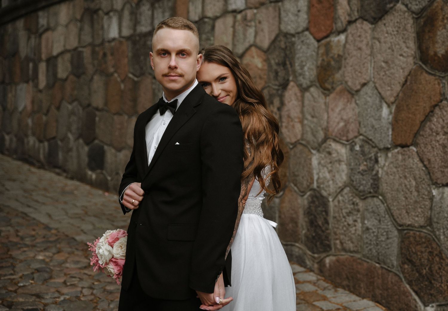 Elegant couple in formal wedding attire pose against rustic stone wall during intimate outdoor ceremony.