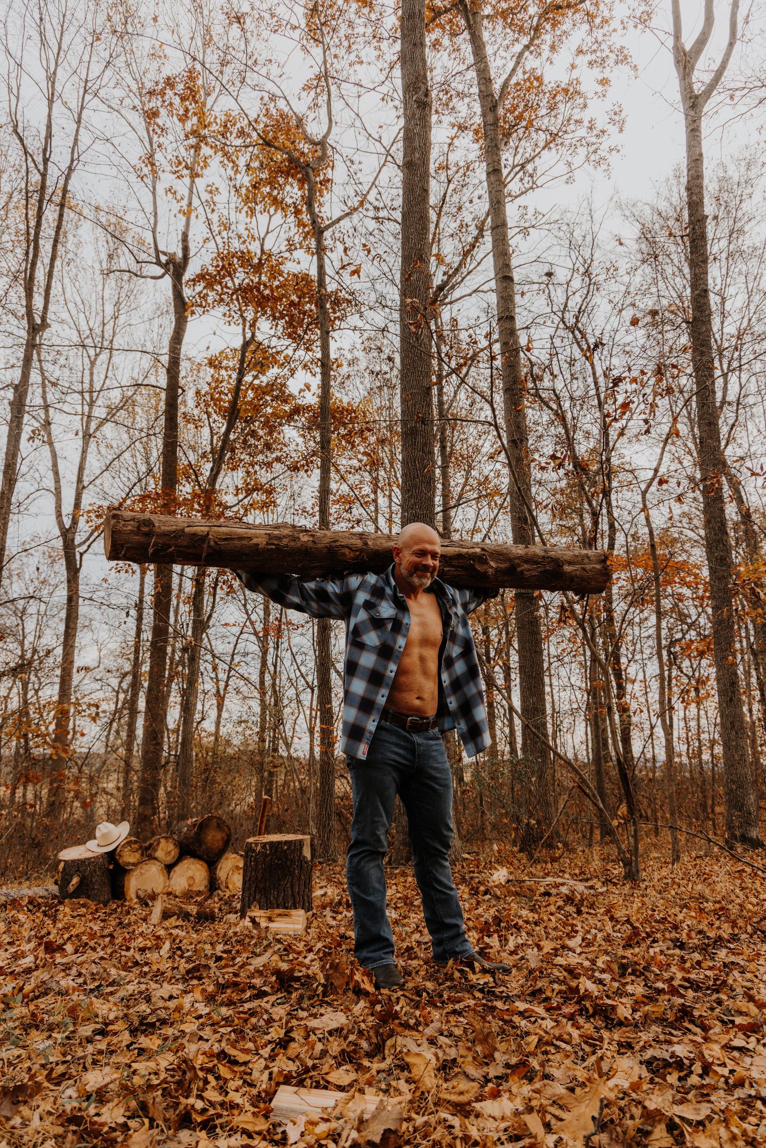 Person carrying a large log through autumn forest with fallen leaves scattered on ground in sepia tones.