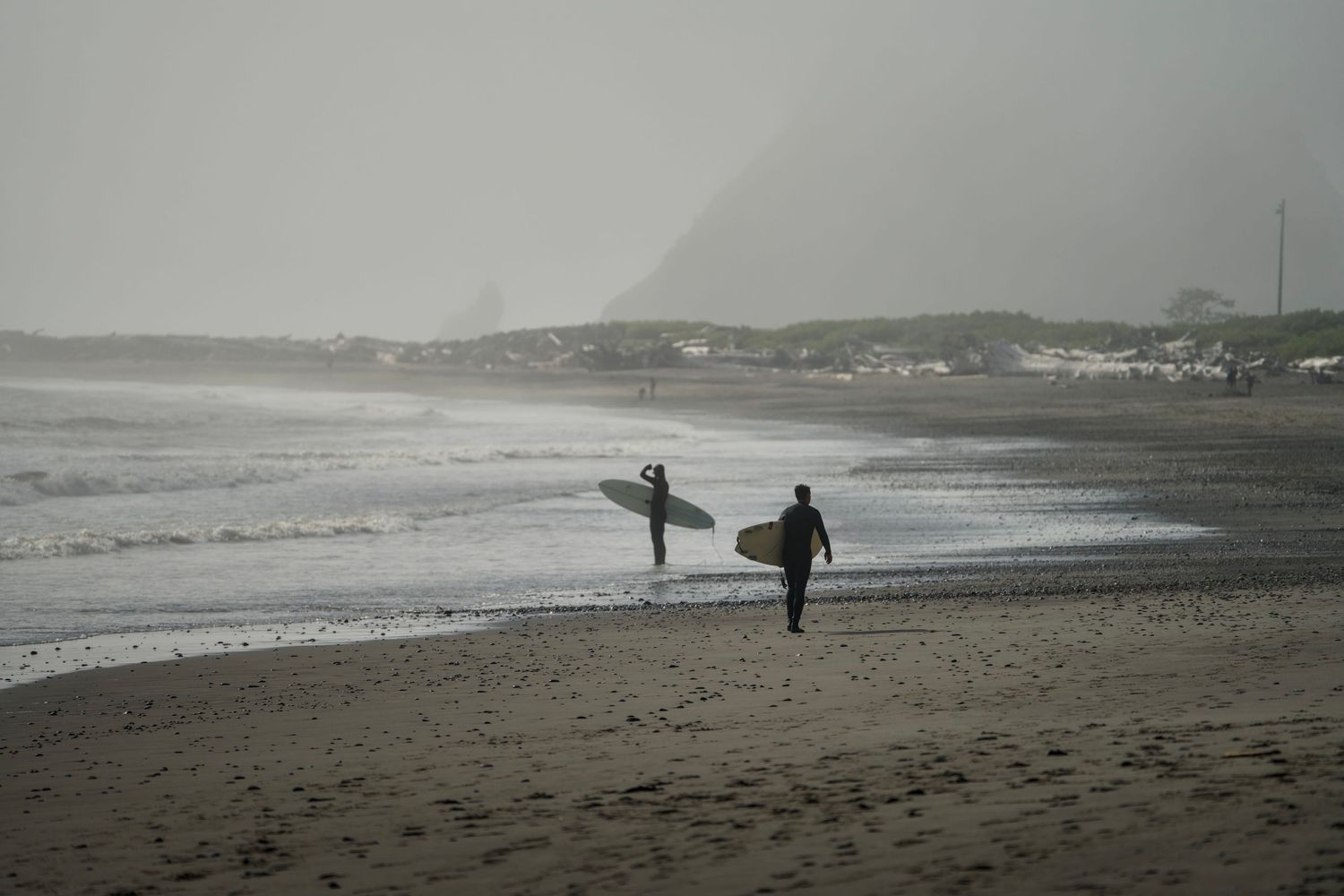 Two surfers carrying boards walk along a foggy, dark sand beach toward a hazy, mountainous coastline.