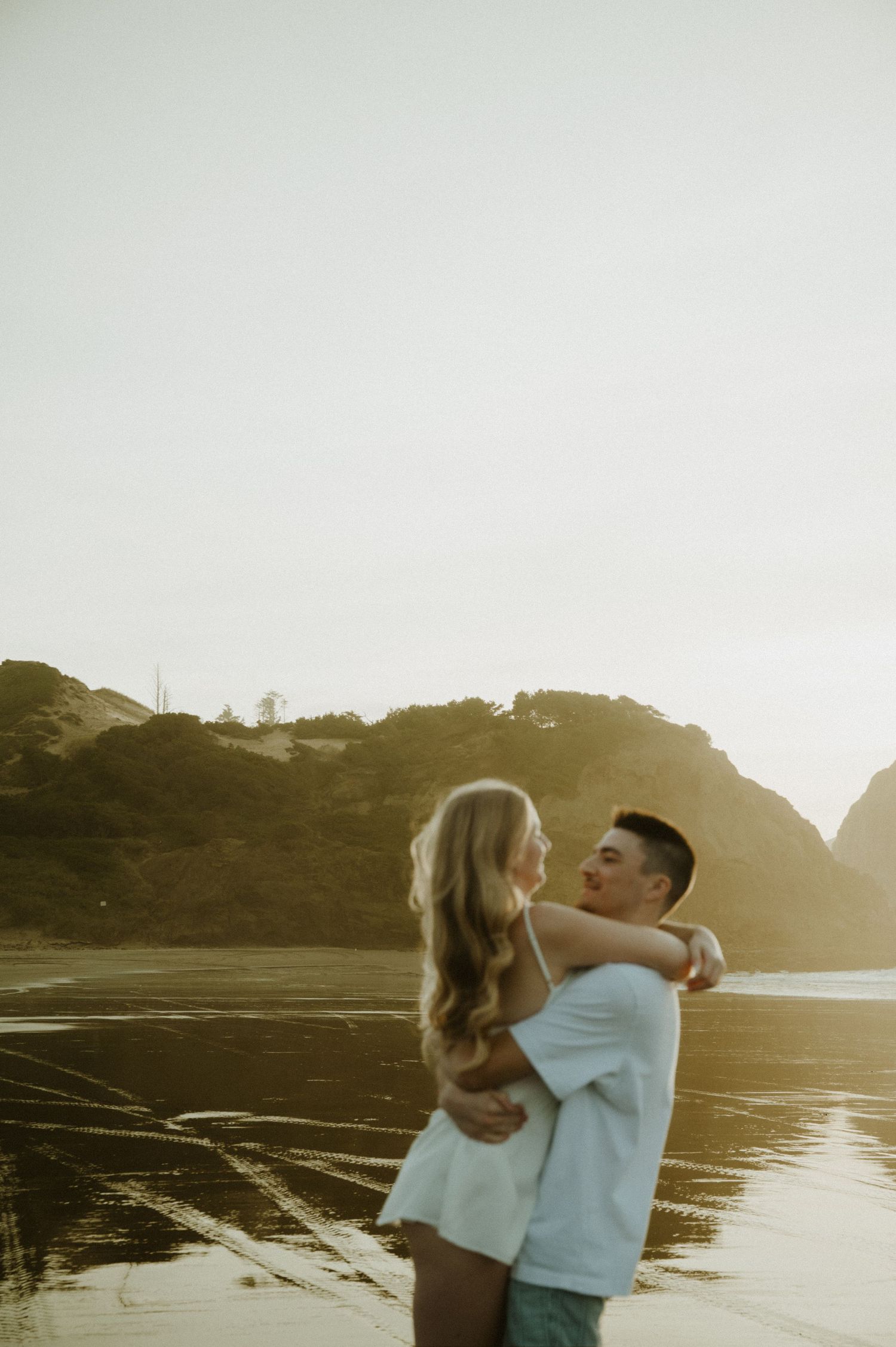 Couple shares a tender moment on the beach during sunset with coastal cliffs silhouetted behind them.