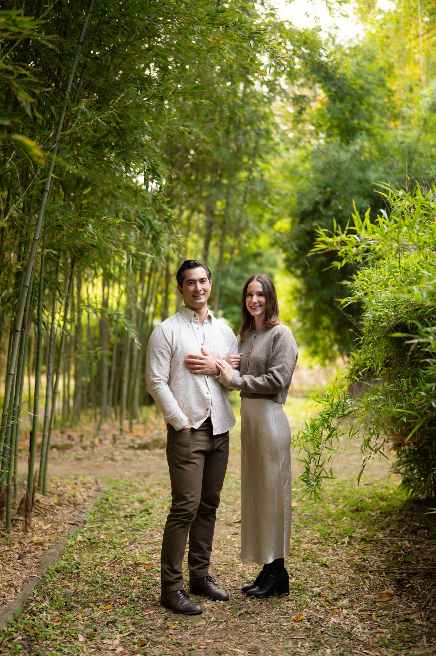 Couple posing together on a bamboo forest path, woman in grey dress embracing man in white shirt.