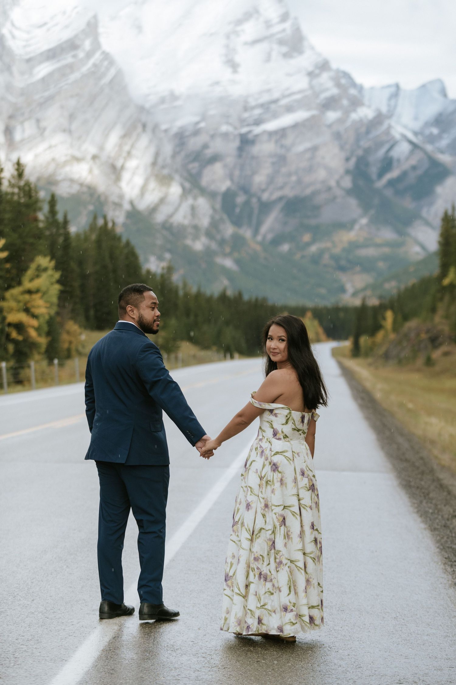A couple holds hands on an empty mountain road in Banff National Park with snow-capped peaks in the background.