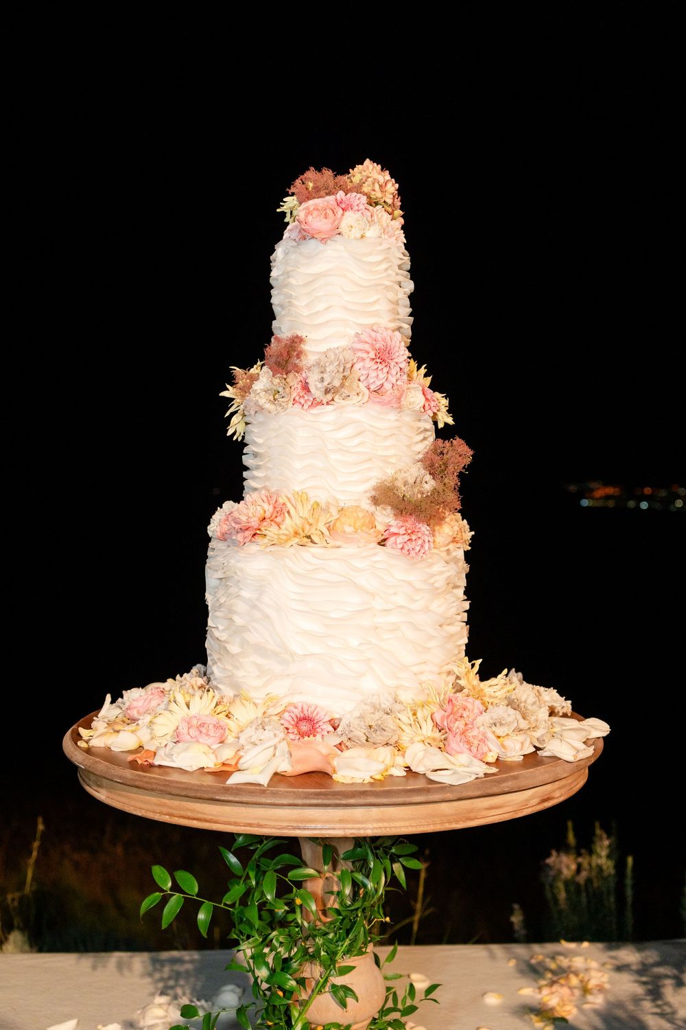 A white three-tiered wedding cake decorated with delicate pink flowers and coconut shavings sits on a rustic wooden stand.