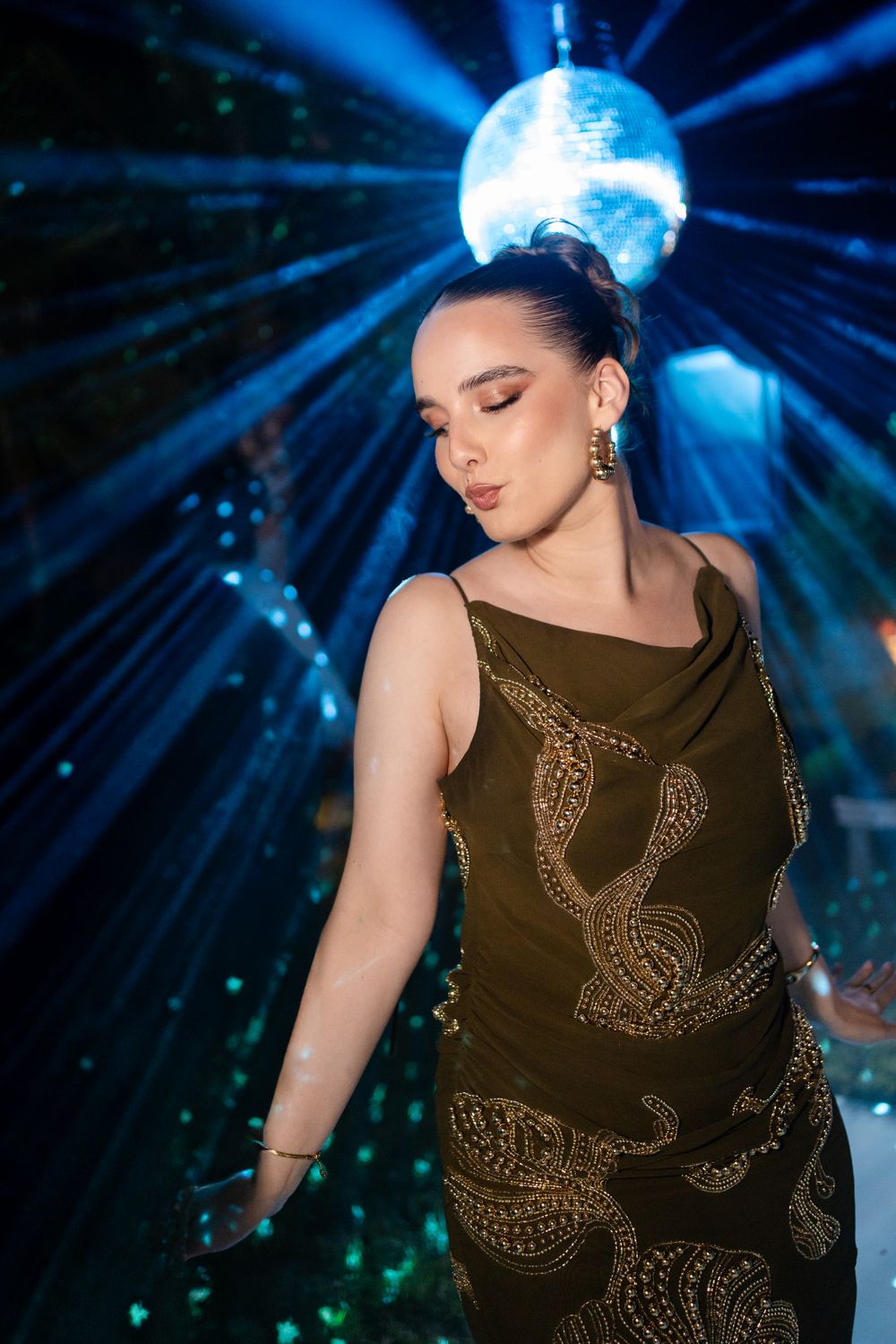 A performer in a brown embellished dress dances under blue disco lights and a glittering mirror ball.