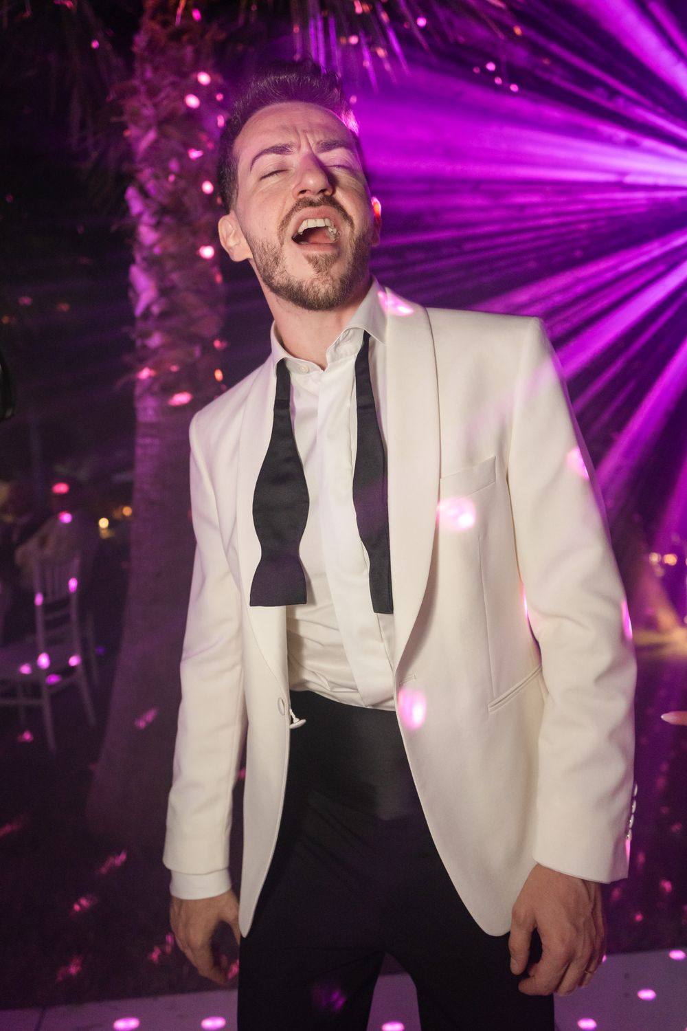A performer in a white tuxedo jacket and black tie laughs joyfully under dramatic purple stage lighting.