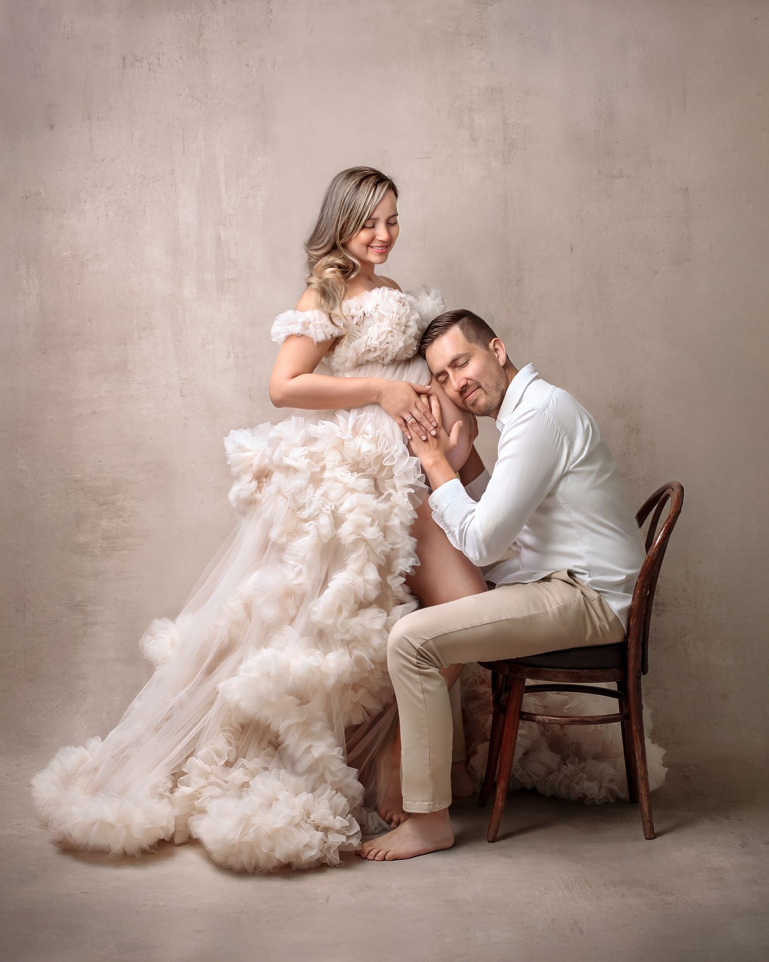 Pregnant woman in a cream couture gown posing standing while her partner sits on a chair in the studio with his head on her belly.