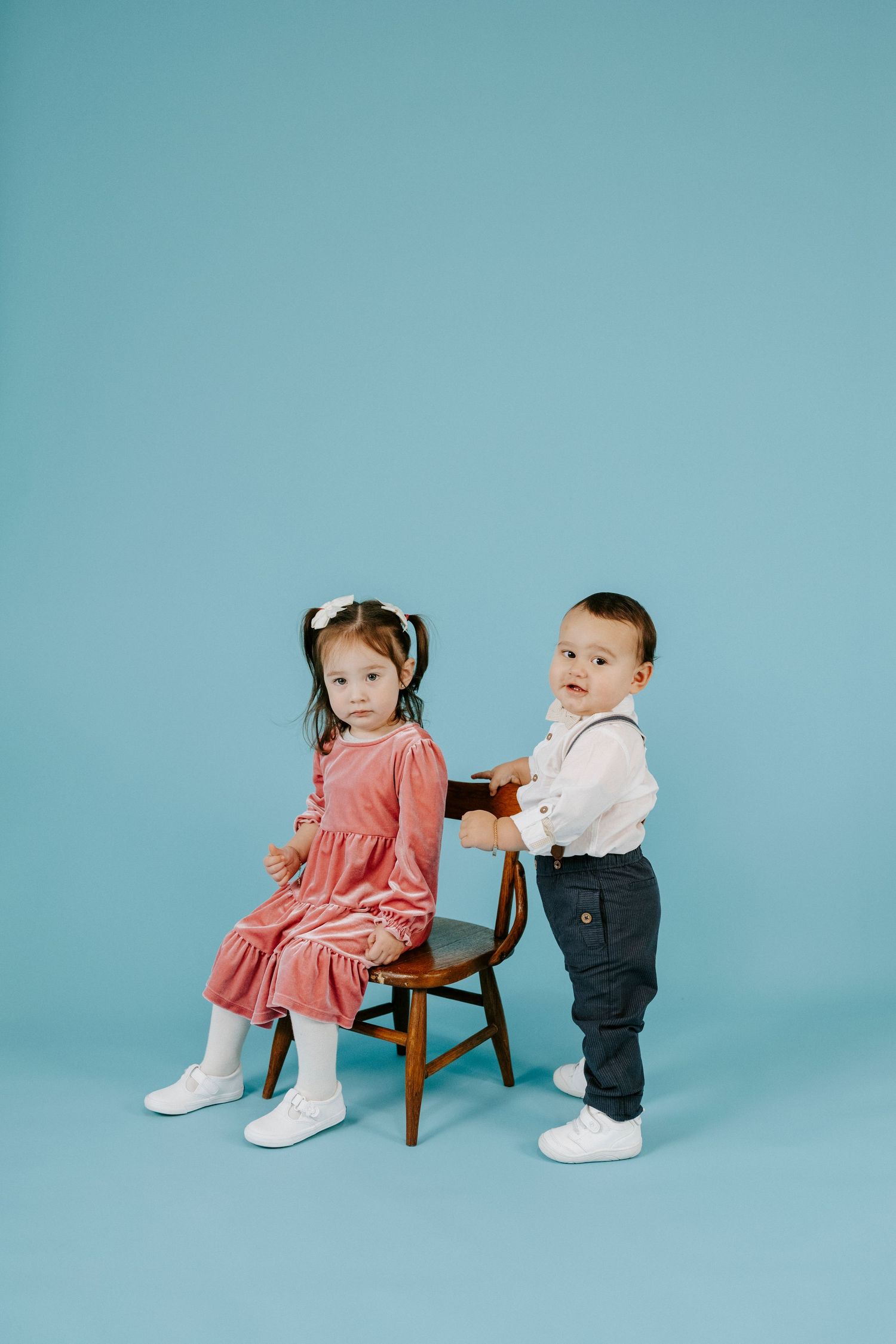 Two toddlers in coordinated outfits interact playfully around a wooden chair against a blue studio backdrop.