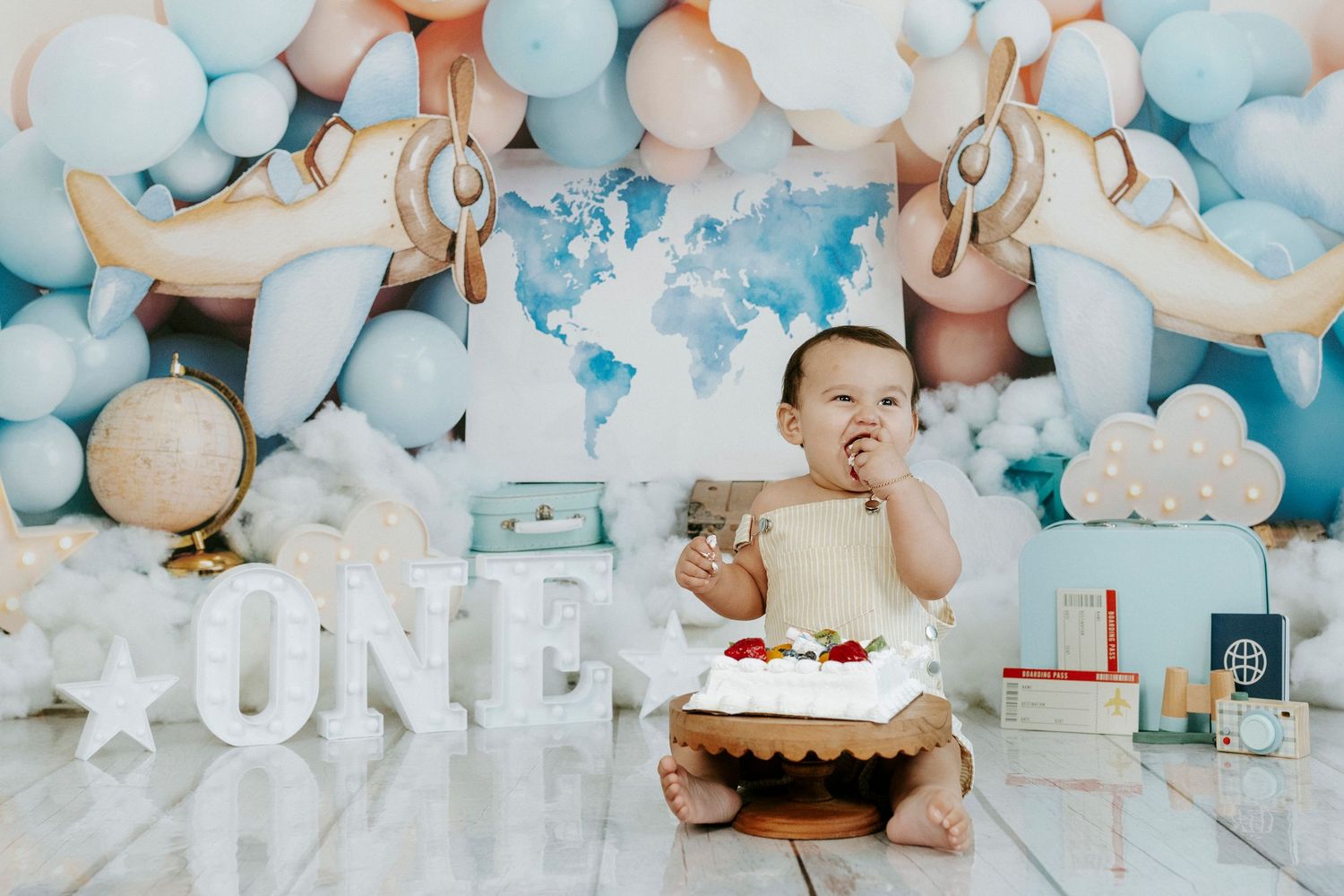 Baby celebrates first birthday with cake and blue balloon decorations against white backdrop.