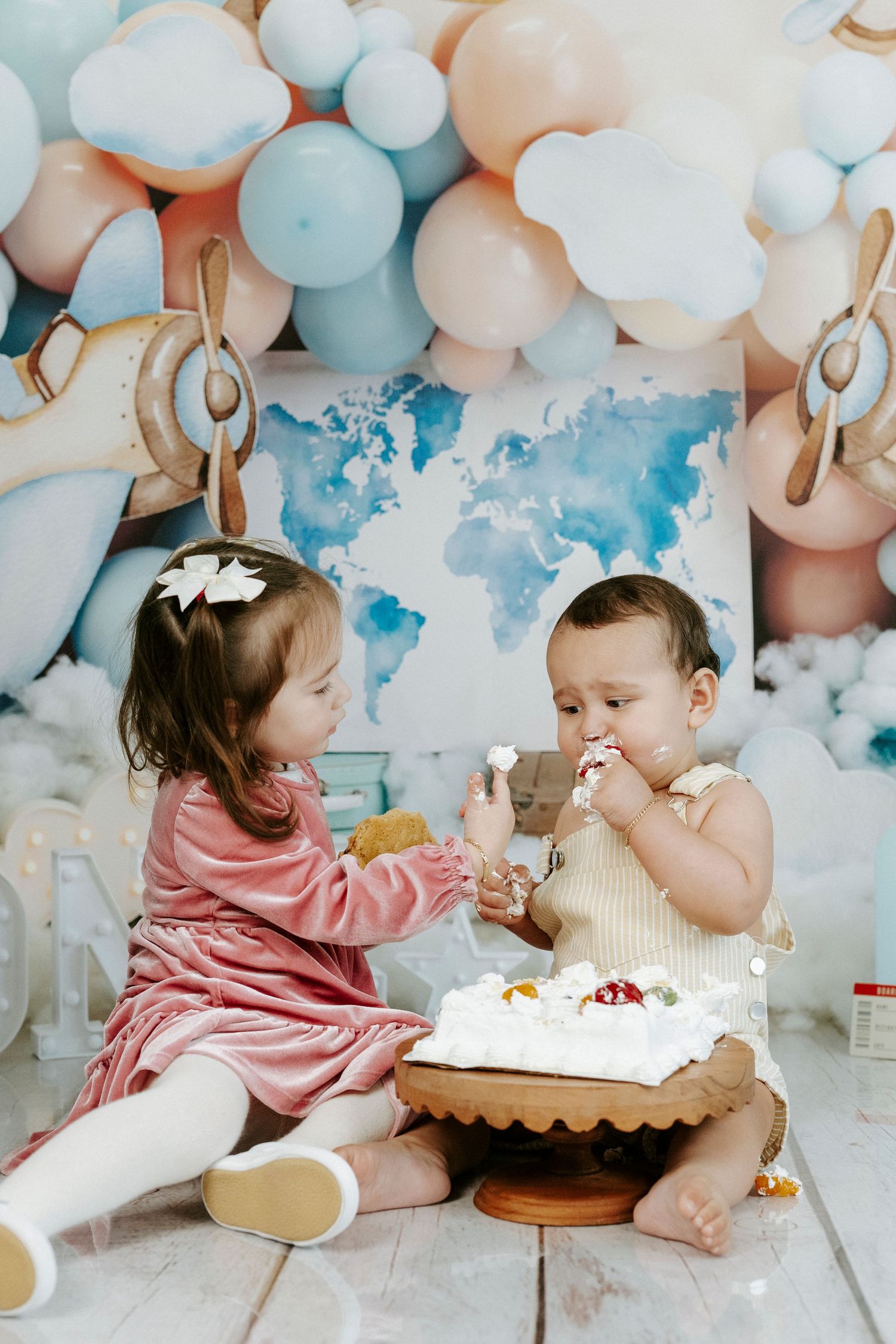 Two babies in pink and white outfits celebrate with cake surrounded by blue and white balloon decorations.