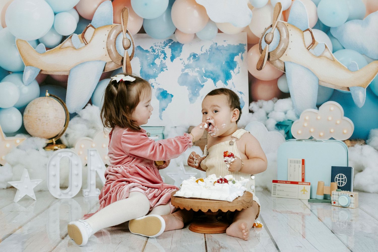 Baby in a white dress celebrates first birthday with cake smash photoshoot surrounded by blue and white balloons.
