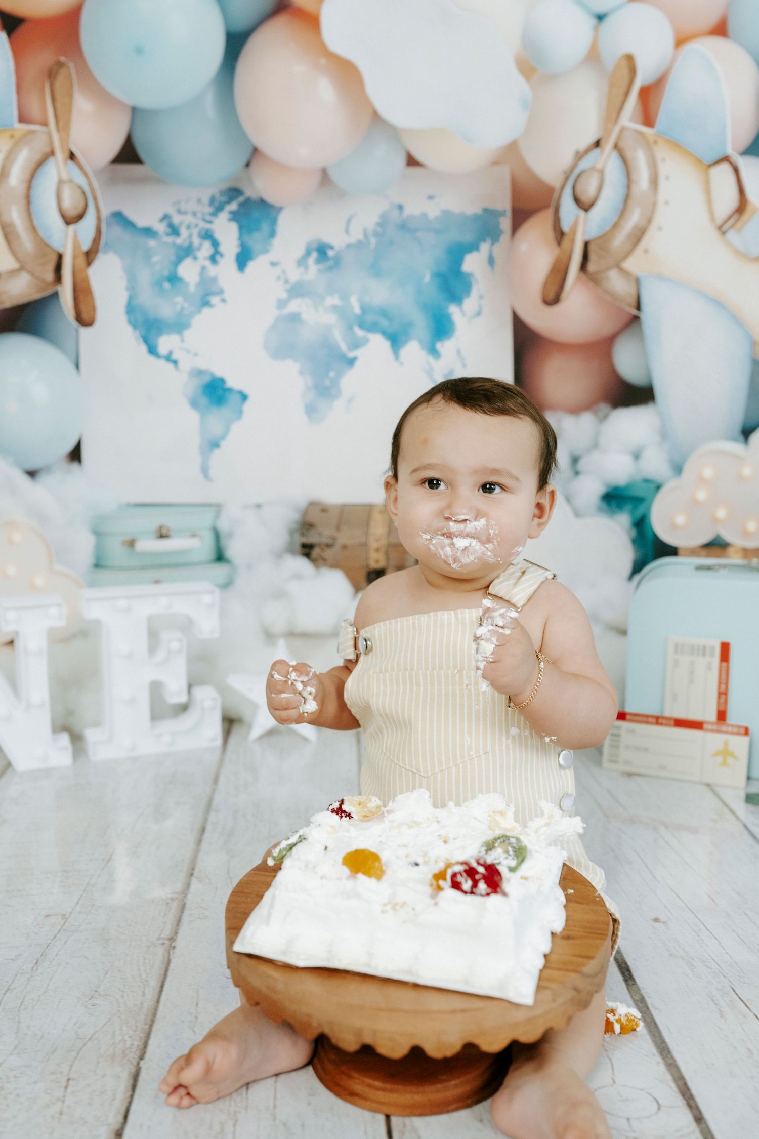 A baby wearing cream colored outfit sits on wooden floor surrounded by pink and blue balloons for cake smash photo session.