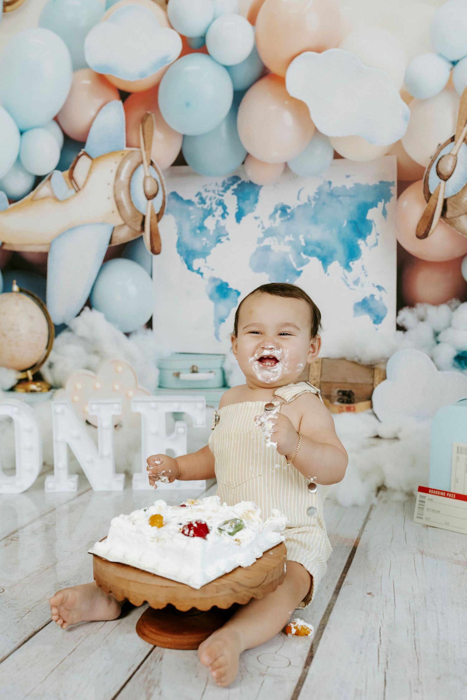 Baby enjoying first birthday cake smash photoshoot with white and blue balloon backdrop on wooden floor.