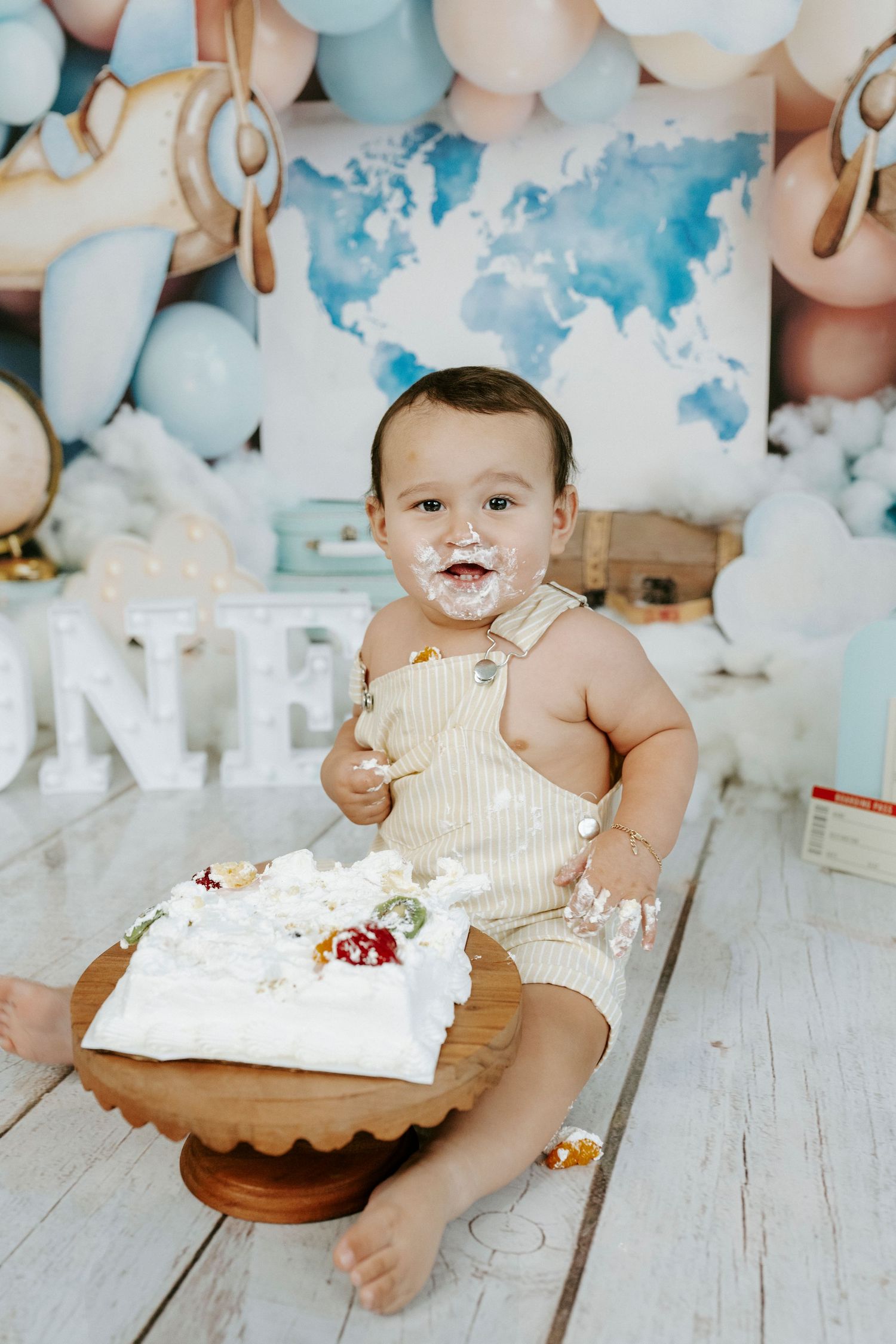 Baby in cream outfit sits on wooden floor surrounded by blue and white balloons during cake smash photo session.