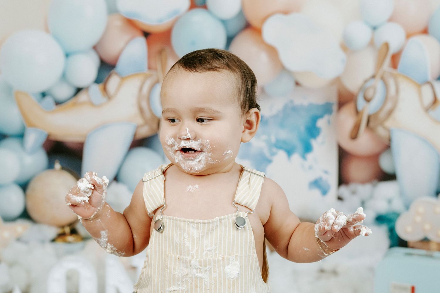 A baby in white sits among pastel blue and white balloon decorations at a first birthday celebration.